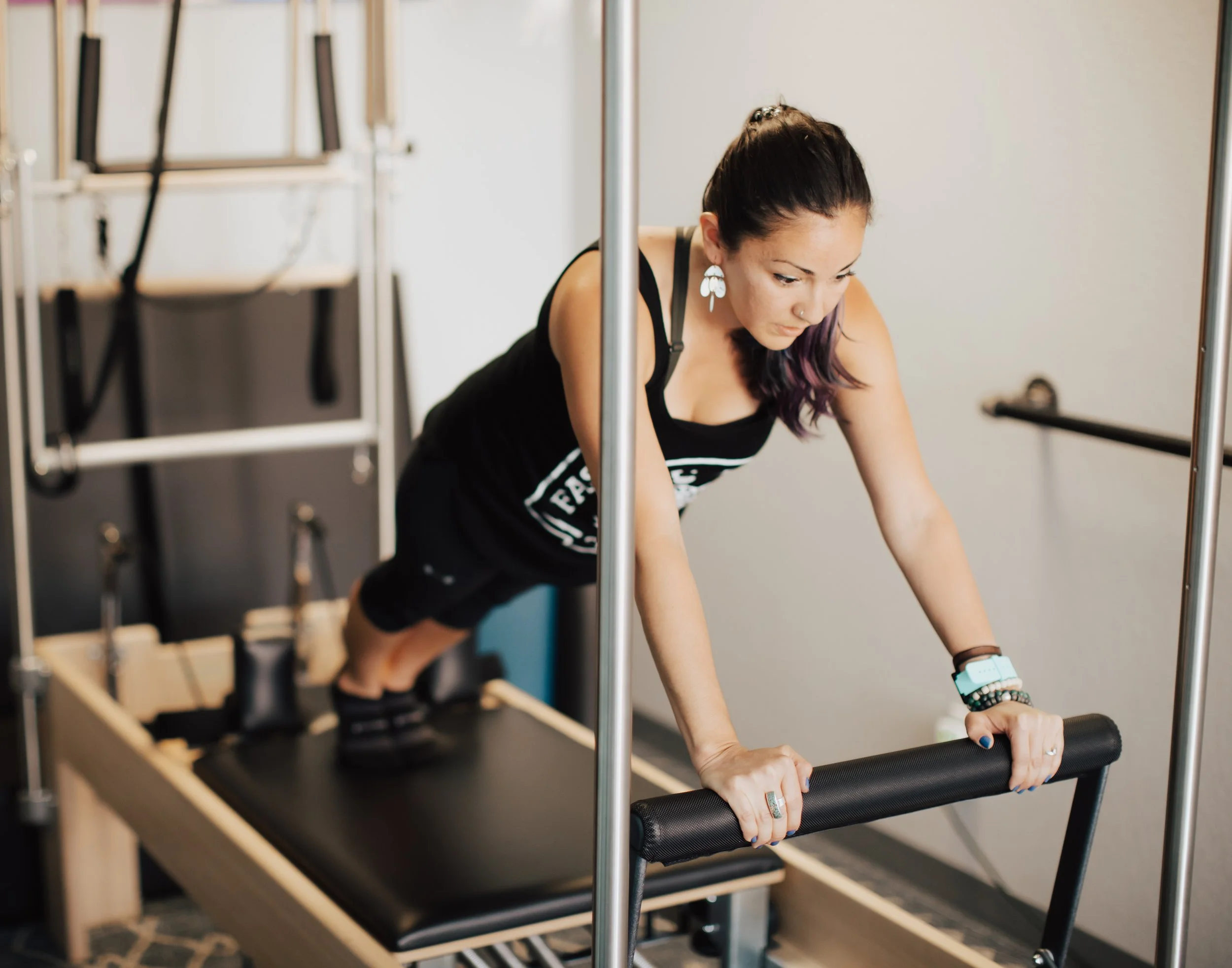 A woman with dark hair tied back, wearing a black tank top and black shorts, doing a plank exercise on a Pilates reformer machine in a fitness studio.