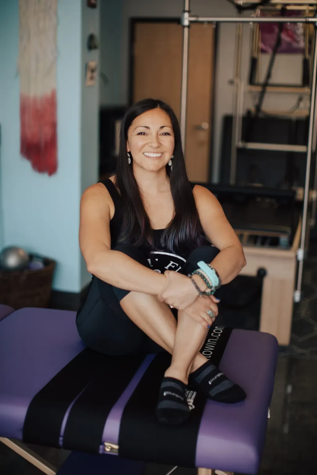 A woman smiling and sitting on a purple and black massage table in a room with fitness equipment and artwork on the wall.