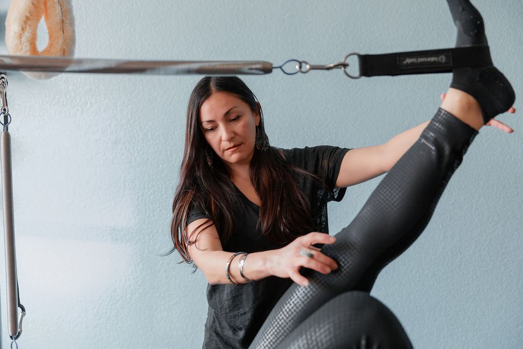 A woman with long dark hair wearing black workout clothes adjusts a client's leg on a Pilates reformer machine.