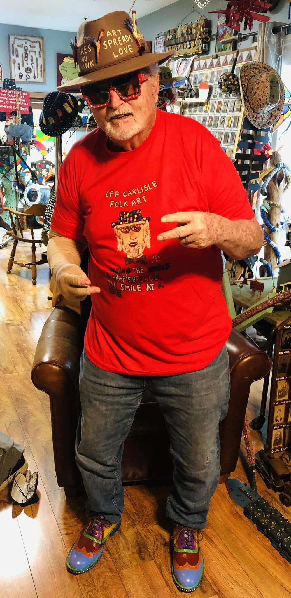 An elderly man with sunglasses, a hat, a red shirt, and colorful shoes making a peace sign inside a gift shop filled with art and craft items.