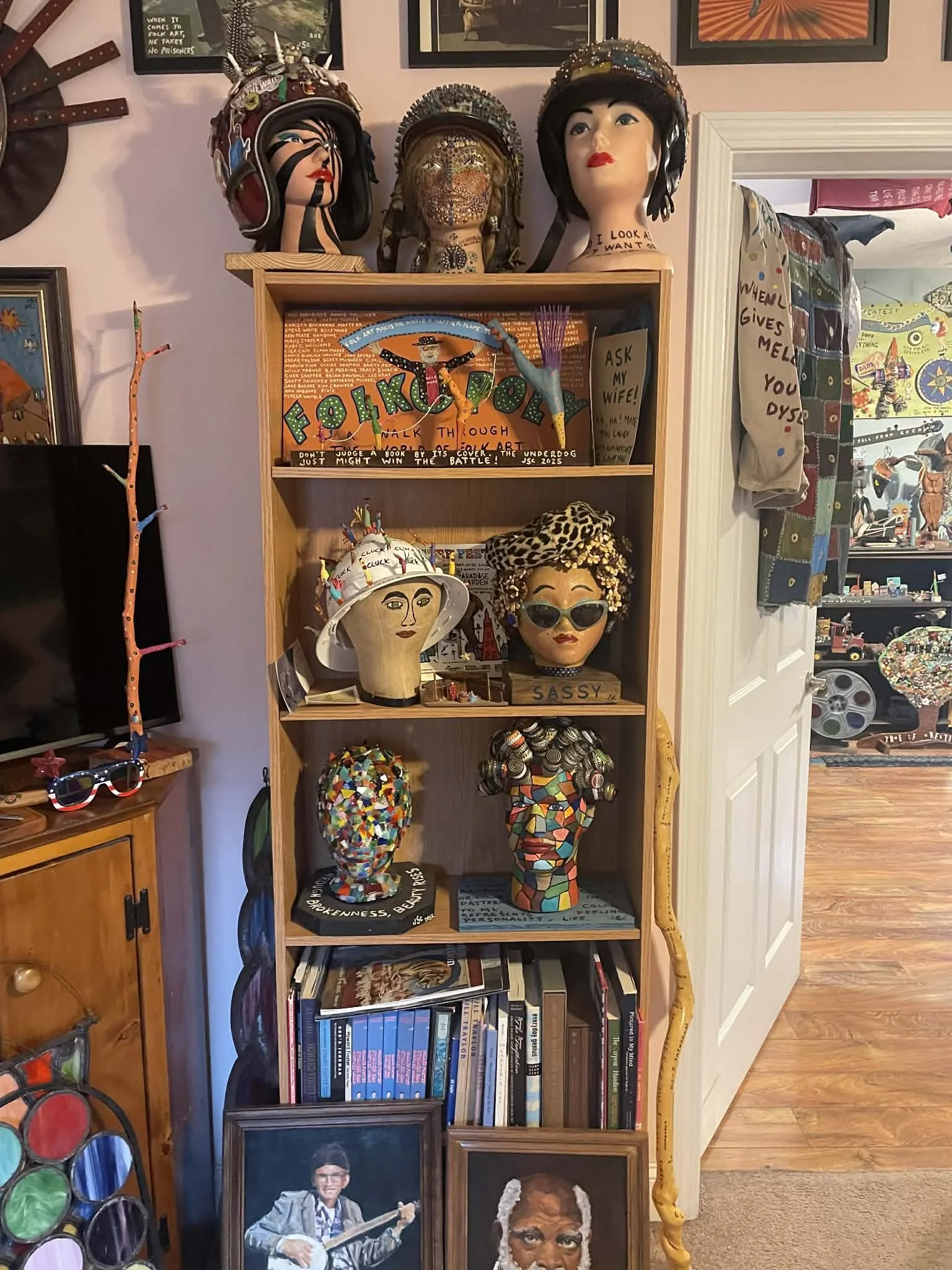 A wooden bookshelf displaying a variety of colorful art mannequins and busts, most with unique hats and accessories, set against a purple wall decorated with framed artwork and signs. Additional handmade crafts and photographs are visible nearby.