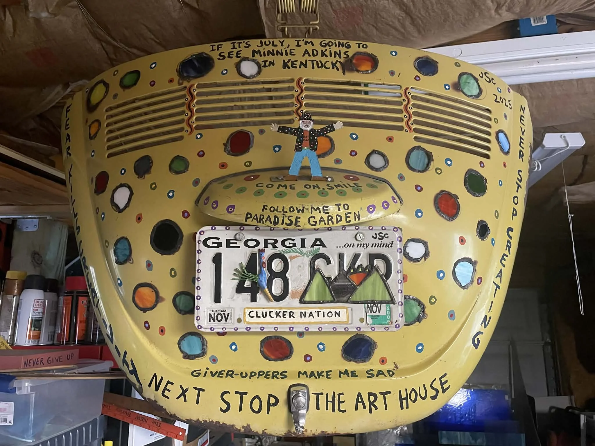 Decorative yellow license plate with colorful circles and handwritten text, featuring a clown figure, and themed around Georgia, Kentucky, and the art house. It includes phrases like 'Follow me to paradise garden' and 'Next stop the art house'.