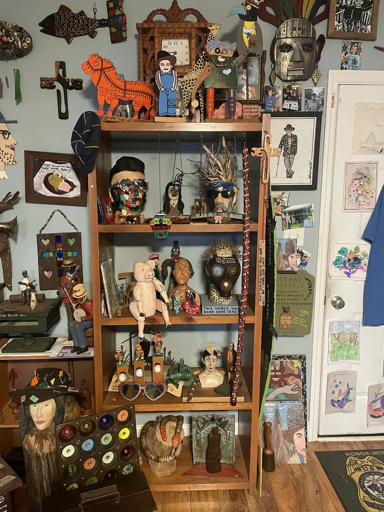 A wooden display shelf filled with various eclectic art pieces, sculptures, and decorative objects, with colorful artwork and photos on the wall around it.