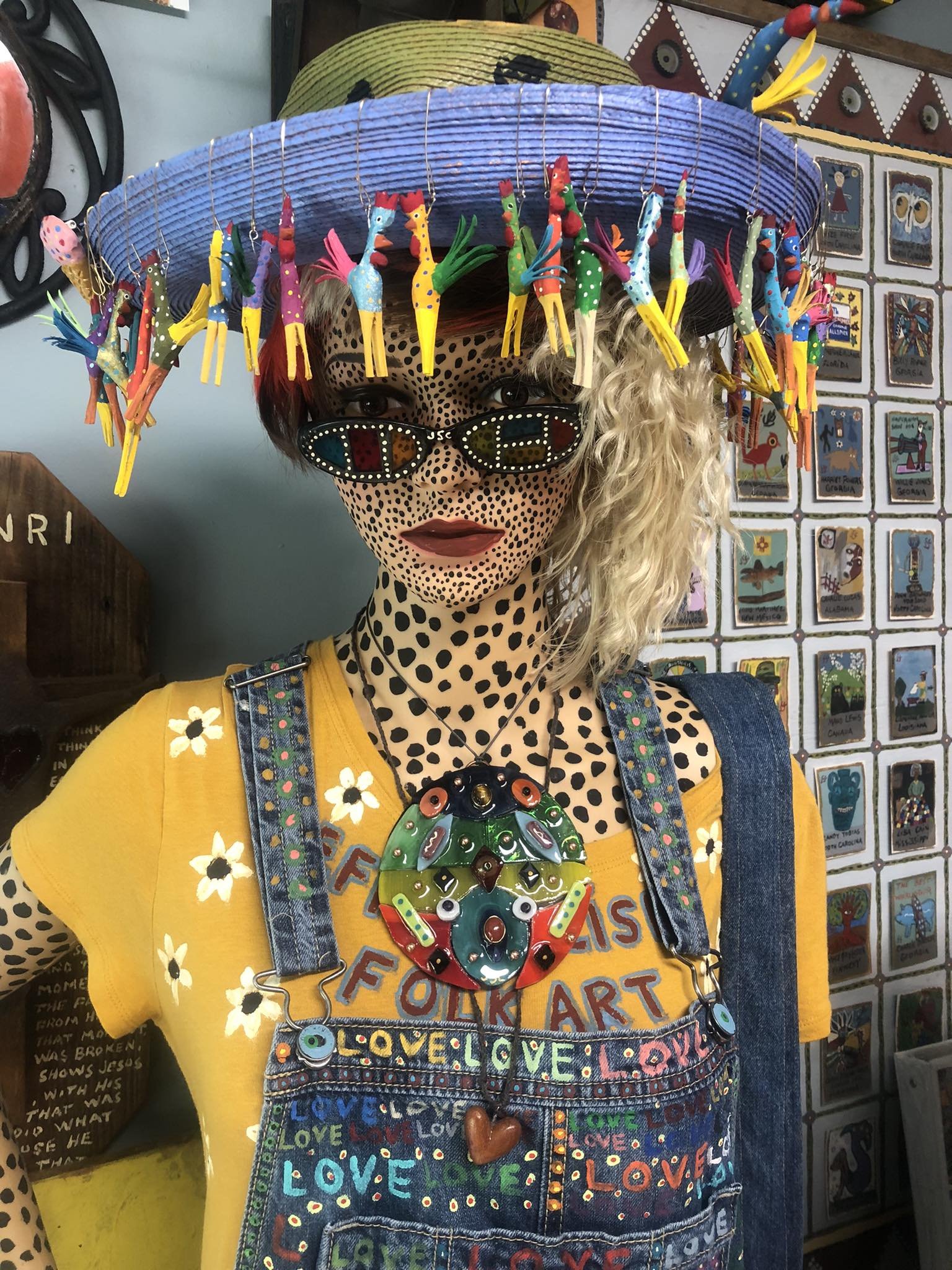A mannequin decorated with a leopard print face and arms, wearing a yellow shirt with flower designs, denim overalls, and colorful accessories. The mannequin has curly blonde hair, decorated with a large colorful necklace, and wears quirky sunglasses