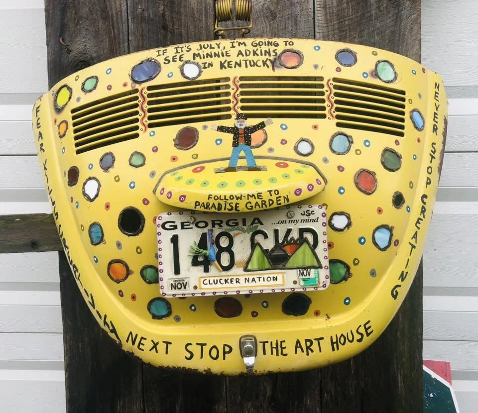 Decorative yellow fiberglass sign shaped like a giant camera with colorful polka dots and text promoting Minnie's Adkins and Paradise Garden, featuring a man with arms outstretched standing on a yellow platform. The sign has a Georgia license plate a