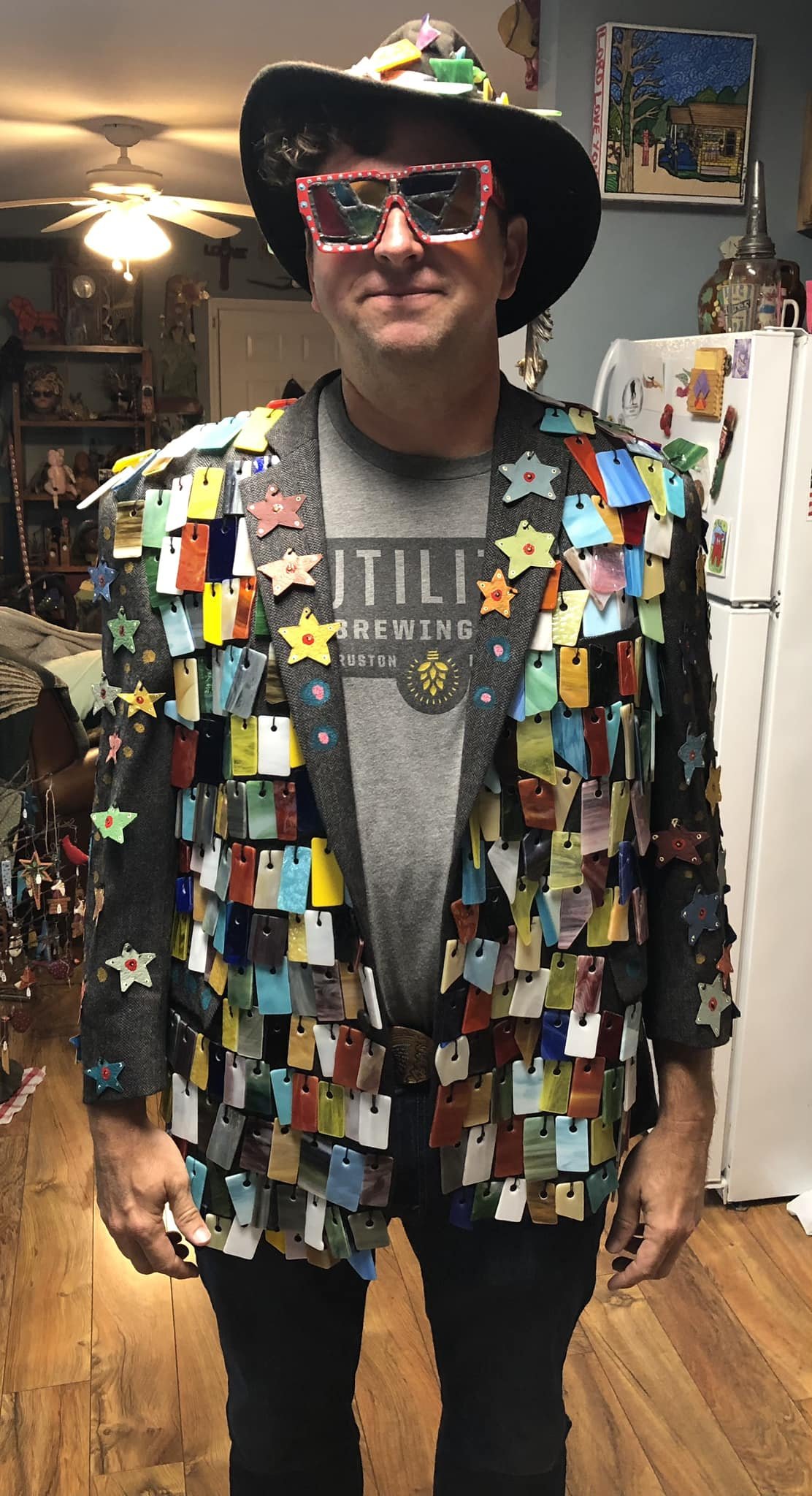 Man wearing a colorful, decorated jacket made of various small, rectangular and star-shaped objects, along with large novelty sunglasses and a wide-brimmed hat decorated with small toys or objects.