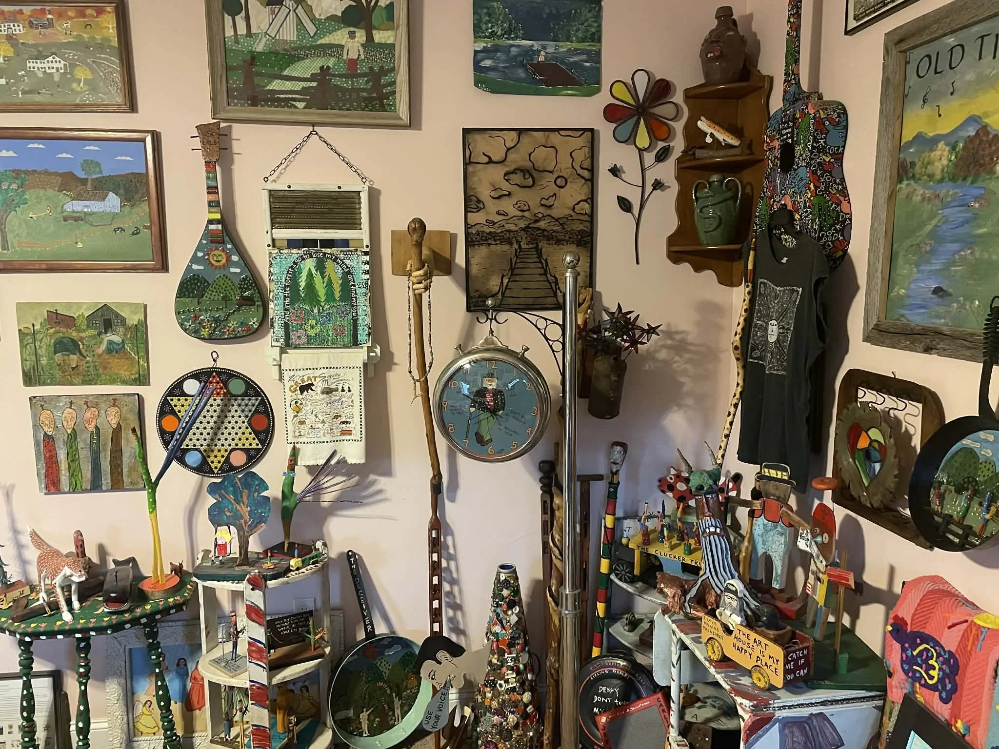 A cluttered room decorated with colorful, folk art style paintings, knickknacks, and various handmade crafts and toys displayed on shelves and tables.