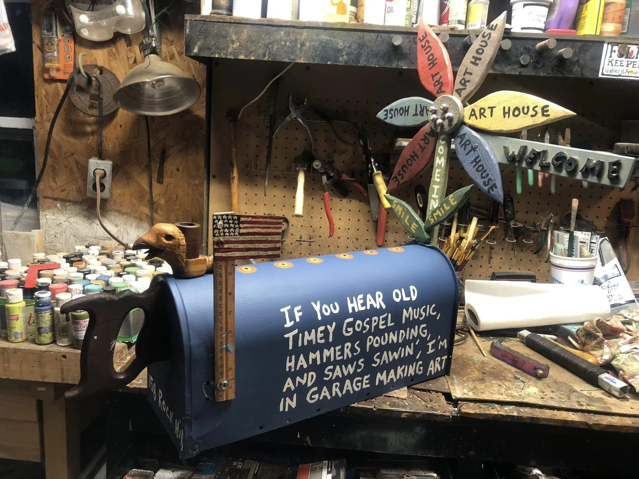 A workshop with a blue mailbox decorated with handwritten white text, surrounded by paint bottles, tools, and a pinwheel with the words 'Art House' and 'Welcome' on its leaves.