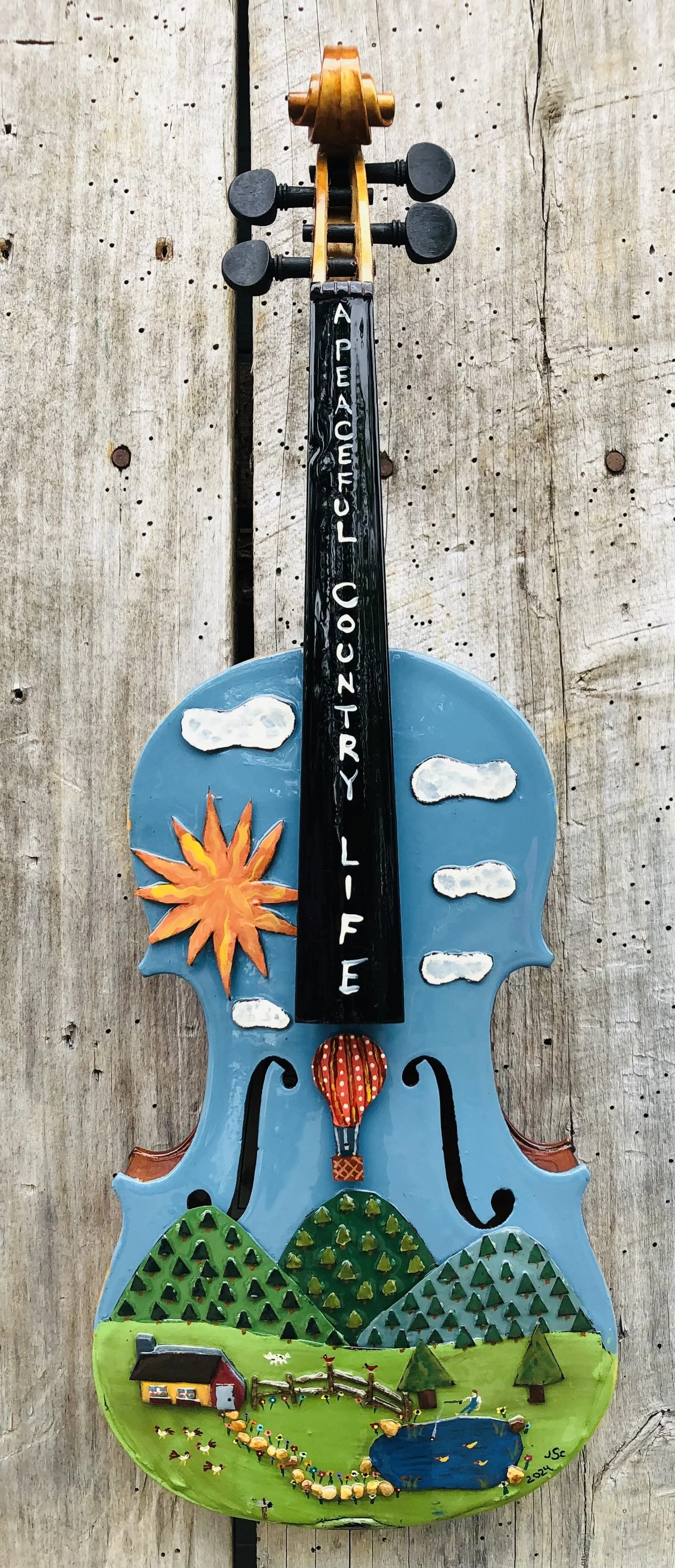 Decorative wall hanging shaped like a violin with a mountain scene, sun, clouds, house, trees, pond, and the phrase 'A Peaceful Country Life' written on it.
