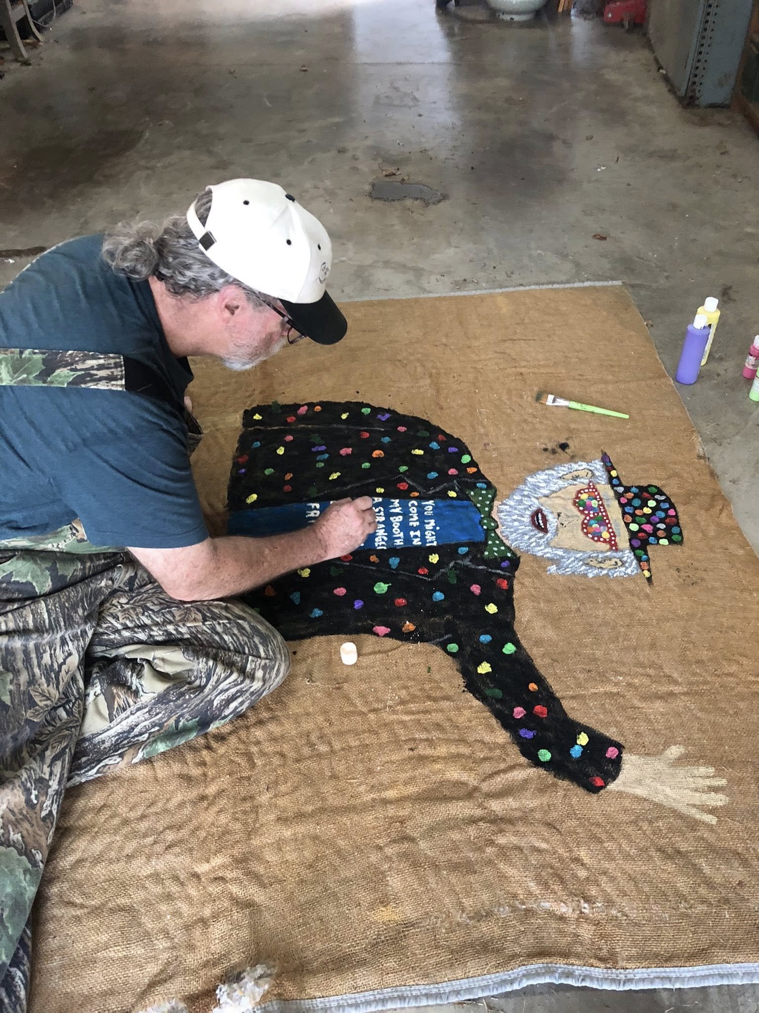 A man working on a colorful fabric art piece in a garage or workshop, laying on the ground and using paint.