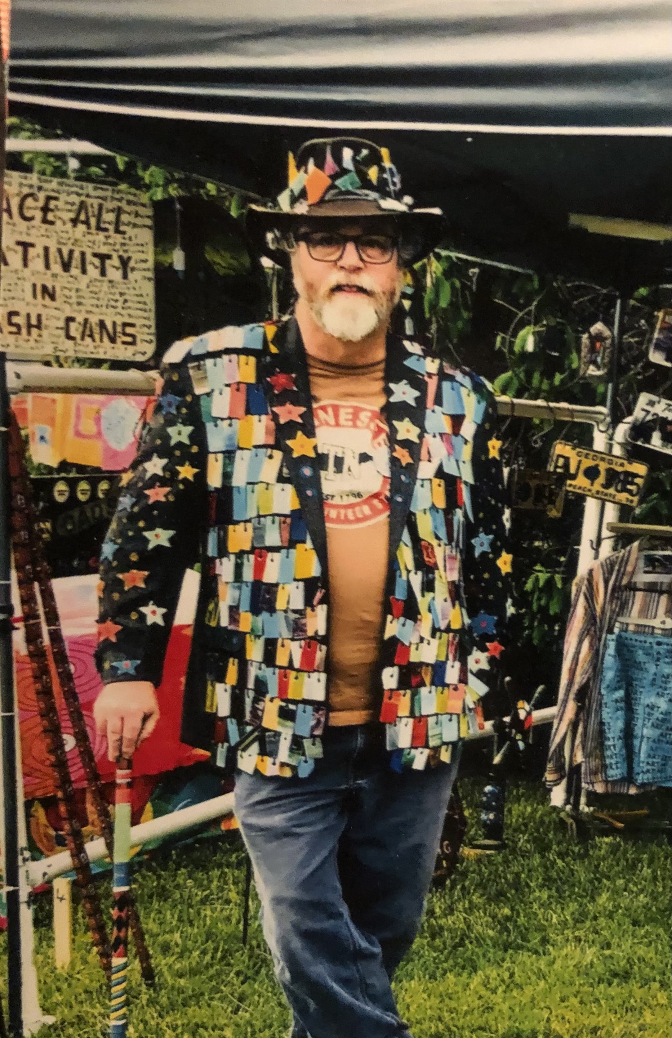 A man dressed in a colorful, star-patterned jacket and hat, standing at an outdoor market stall with signs and clothing items around him.