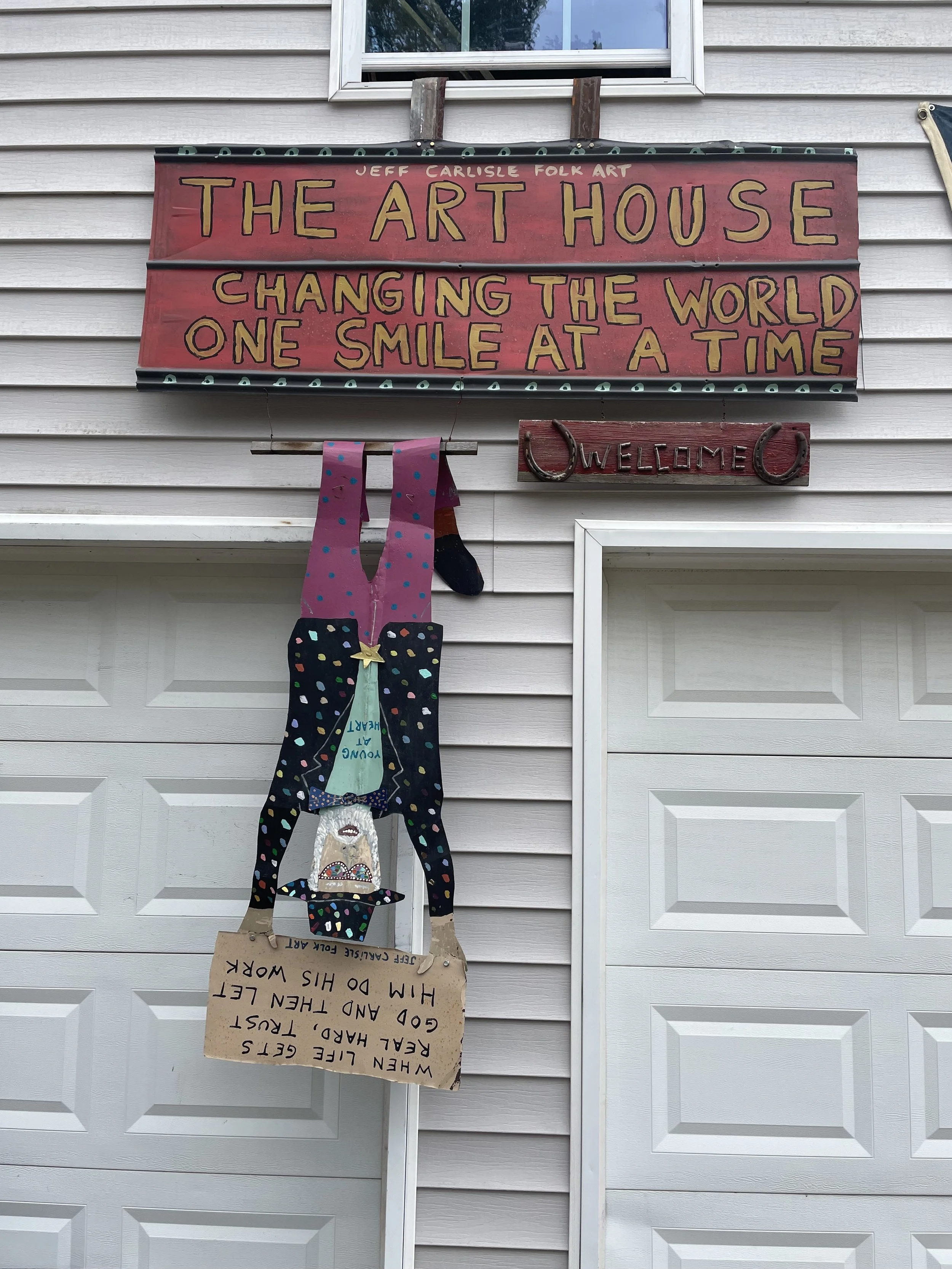 Exterior of a house with signs and artwork. A large red sign with yellow and black lettering reads, 'The Art House - Changing the World One Smile at a Time.' A smaller sign says, 'Welcome.' A hanging paper artwork features a cartoon person upside down, with a message that says, 'When life gets hard and then let go and then let him do his work.'