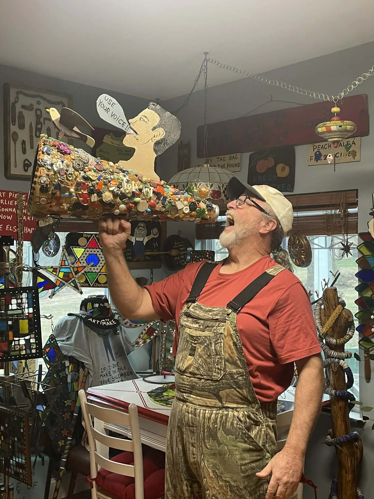 An older man with glasses, a beige cap, a red shirt, and camouflage overalls is laughing and holding a decorated piano-shaped object with buttons and jewelry, in a room filled with colorful art and decorations.