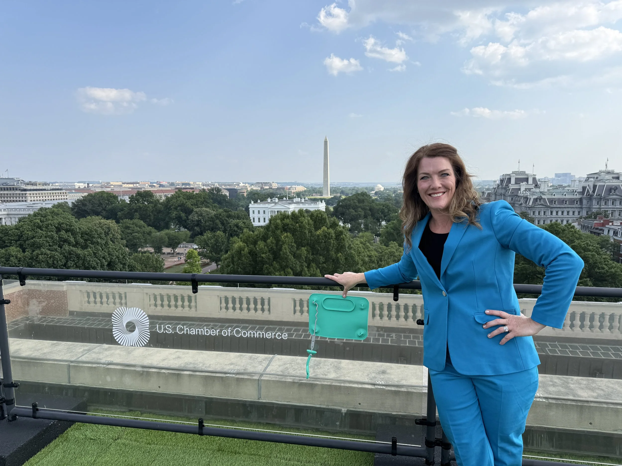 Beth Benike, in a bright blue suit, smiling and displaying her invention, the Busy Baby Mat, on a rooftop with a view of the White House in Washington, D.C., including the Washington Monument in the background.