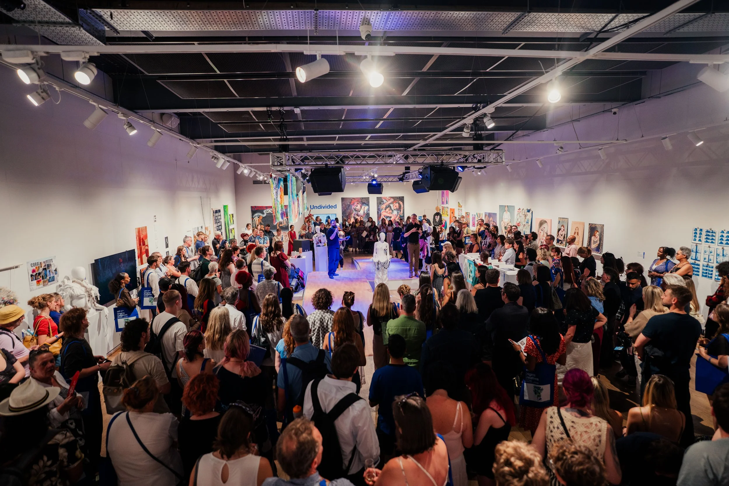 Large art gallery exhibition with many visitors observing artwork and a speech by Calum Hall. The walls display colourful paintings and mixed media art pieces, and a stage area with bright lighting.