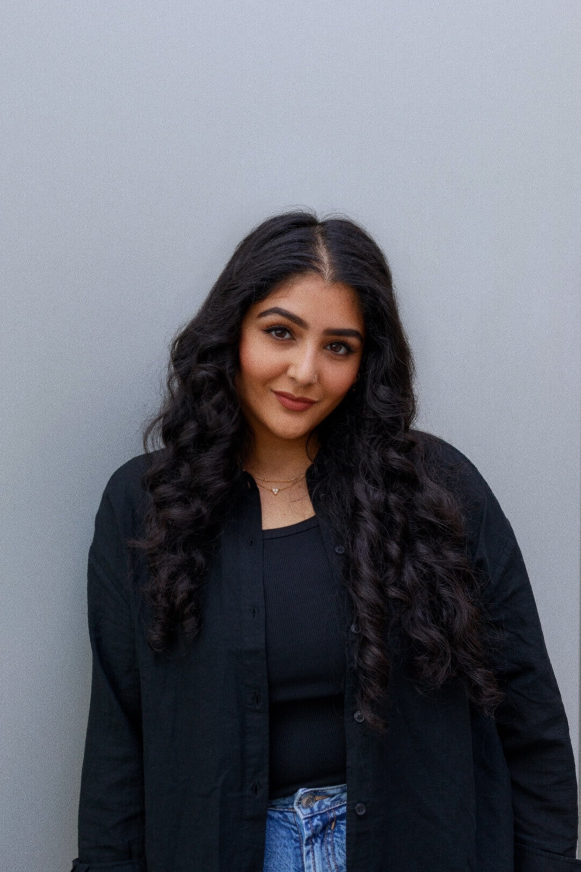 A girl with long curly hair standing in front of a grey wall wearing a black shirt and blue jeans