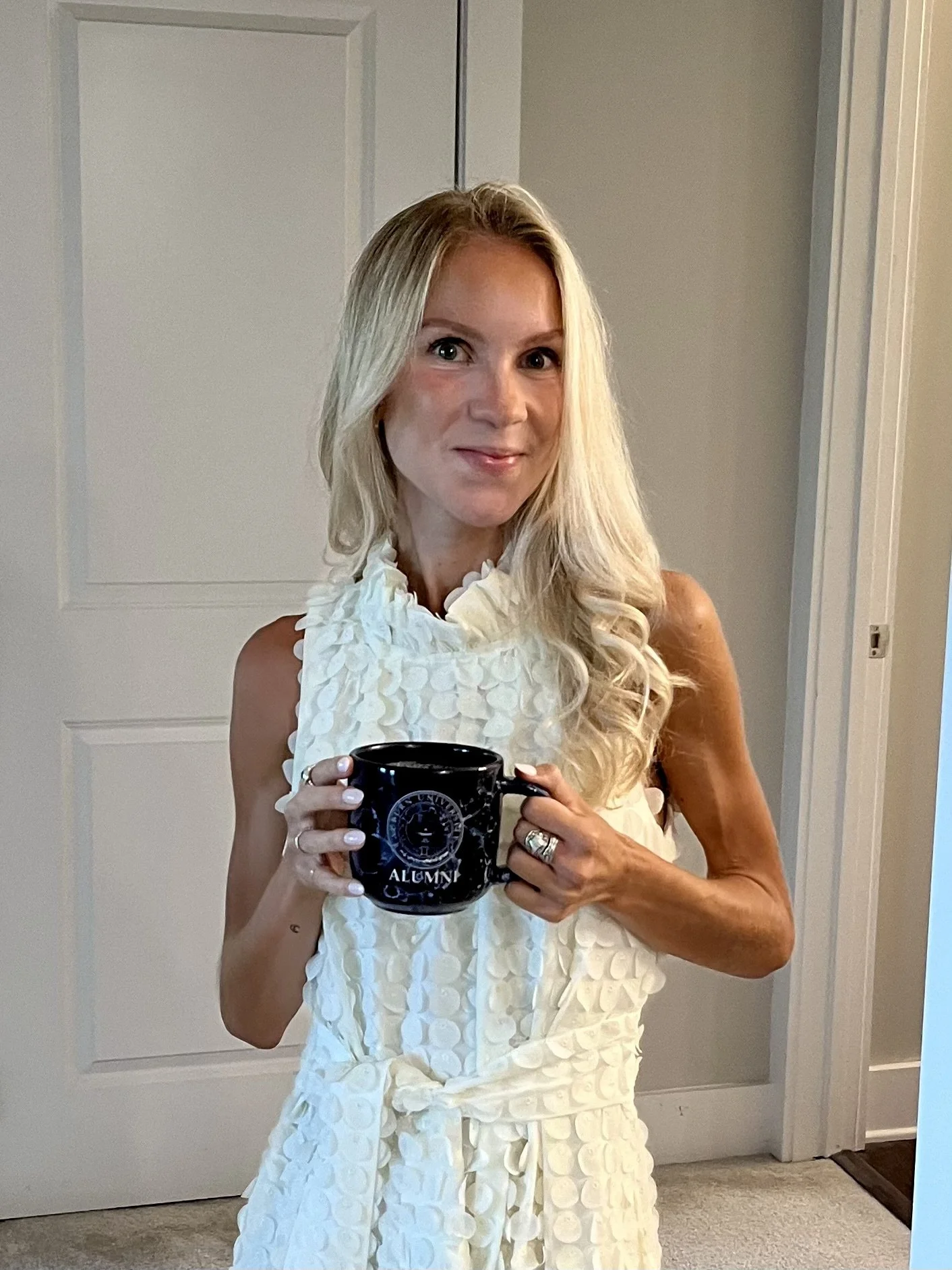 A woman with long blonde hair wearing a sleeveless white dress holding a black coffee mug that has 'ALUMNI' written on it, standing in a room with closed white door.