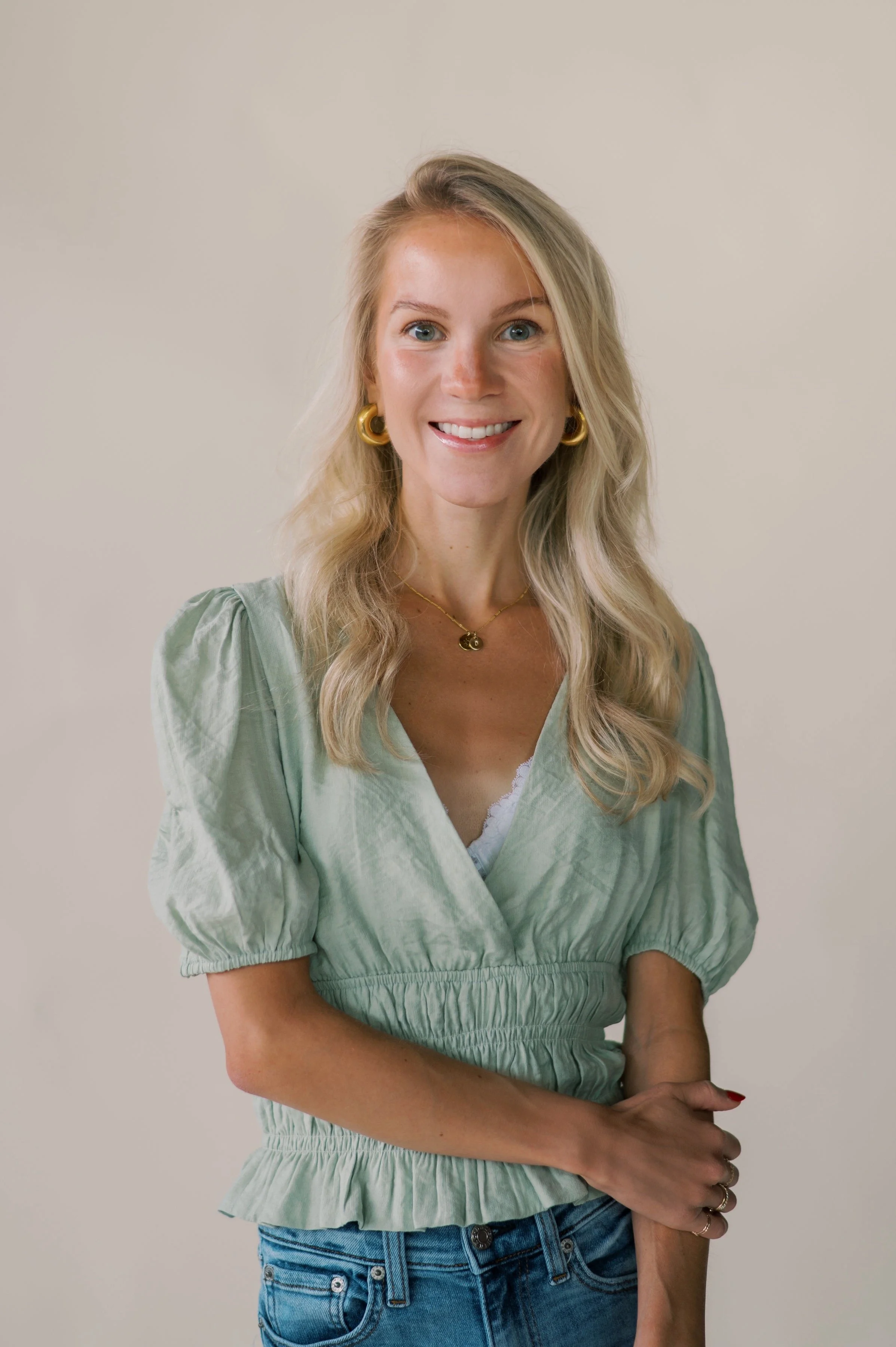 A woman with blonde hair wearing a light green top with puffed sleeves, blue jeans, gold jewelry, smiling against a plain background.