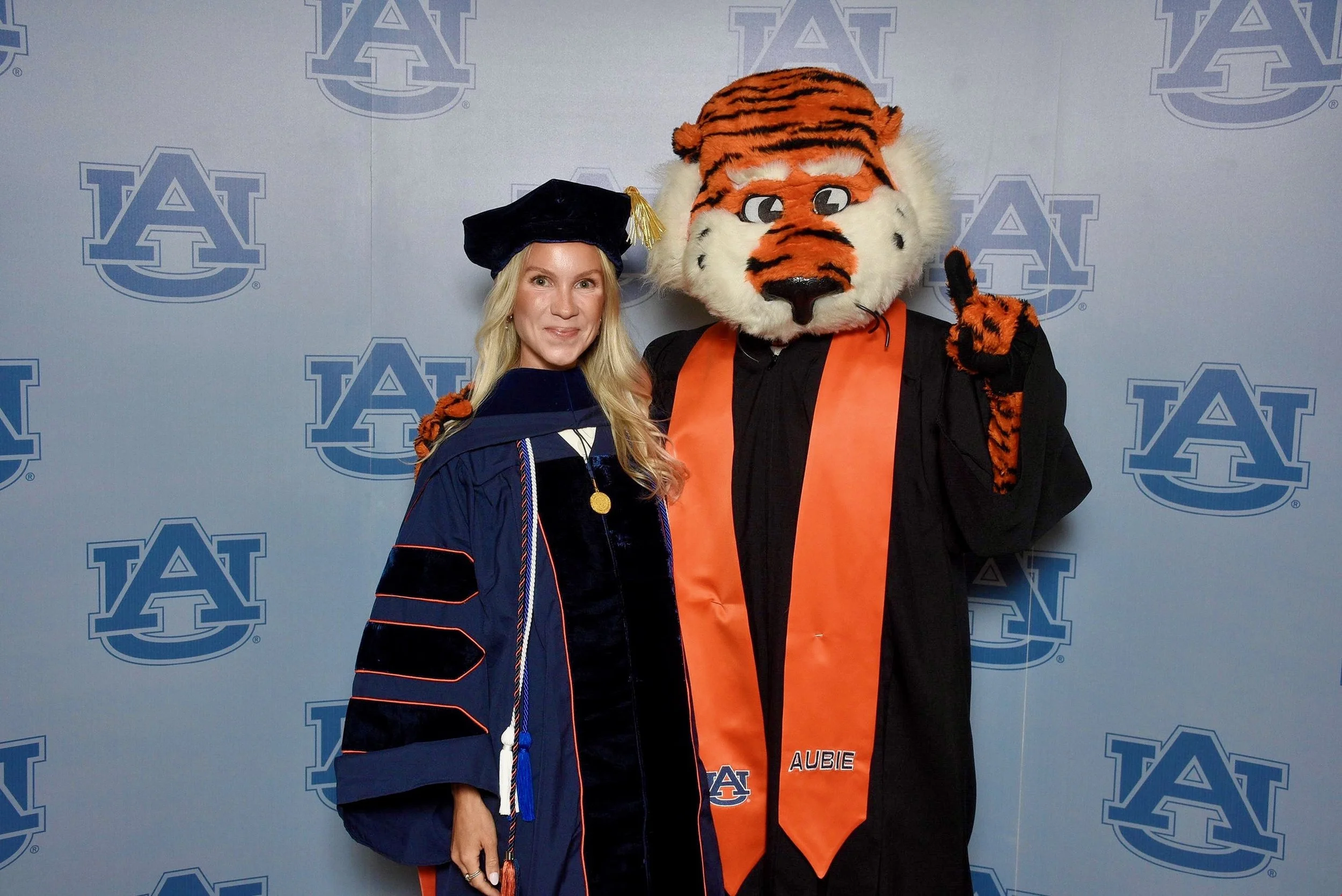 A woman in a graduation gown and cap standing next to a person in a tiger mascot costume, both posing in front of a backdrop with the Auburn University logo.
