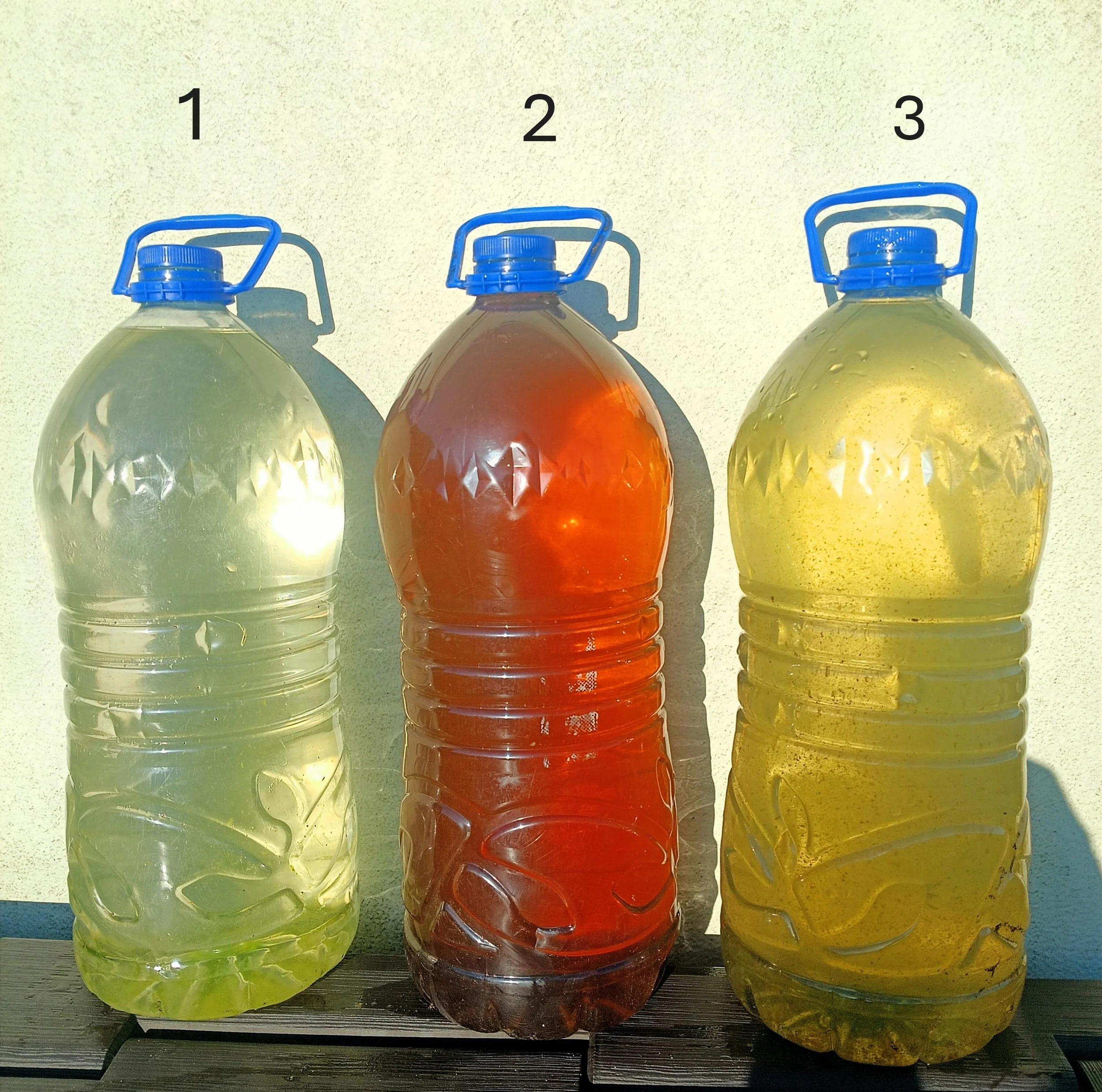 Three large plastic bottles filled with different colored liquids are placed on a wooden surface against a light-colored wall. The bottles have blue handles and caps, and each bottle contains a different colored liquid: clear, red, and yellow.