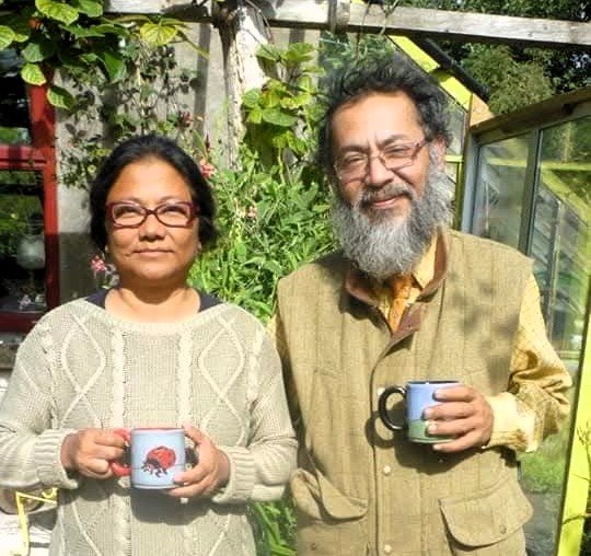 A woman and a man standing outdoors, holding mugs, surrounded by green plants and trees, with sunlight shining.