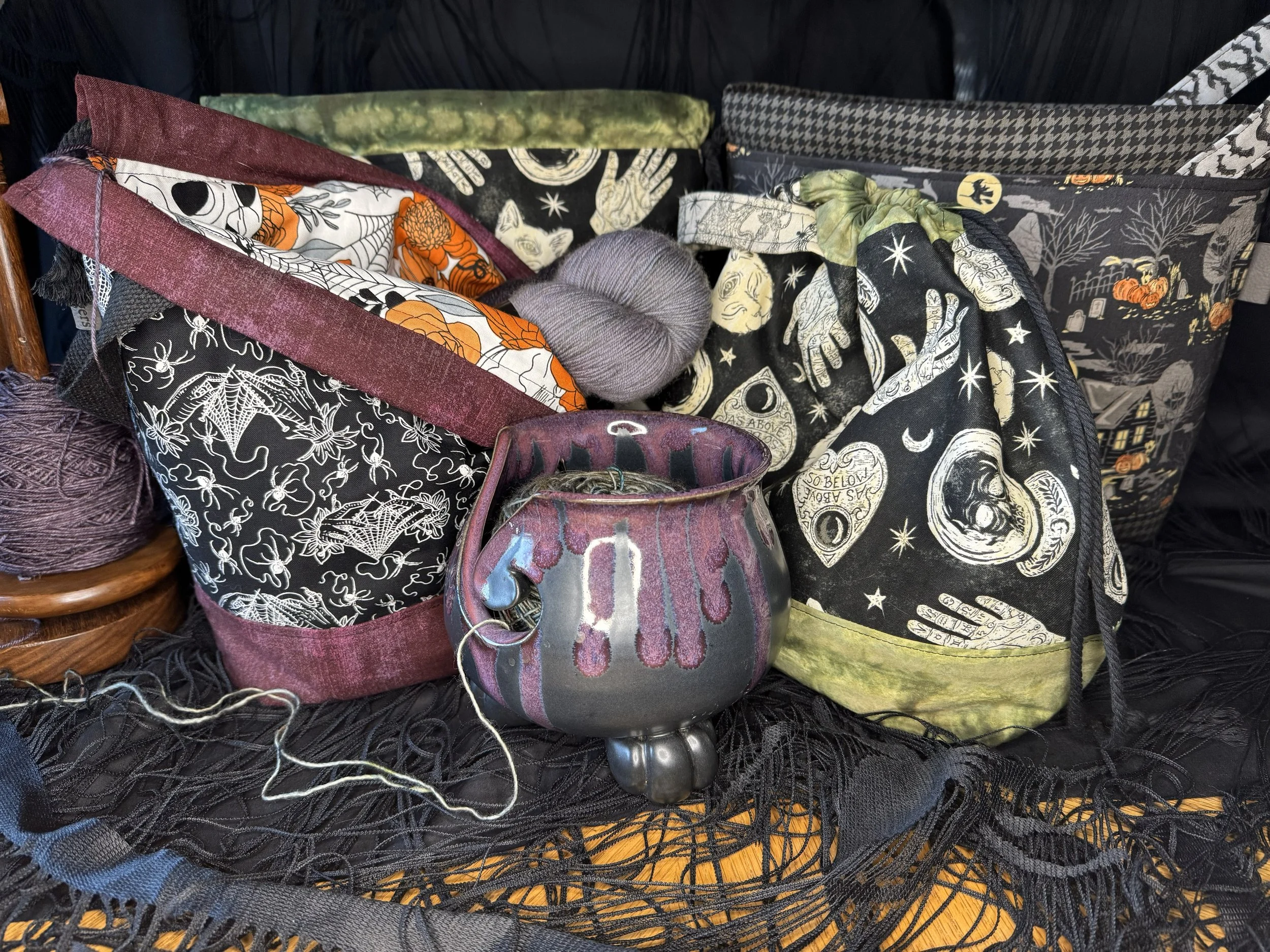 A collection of Halloween-themed tote bags with various spooky designs, a gray ball of yarn, and a small black ceramic cauldron with purple and blue drips, all displayed on black lace fabric.