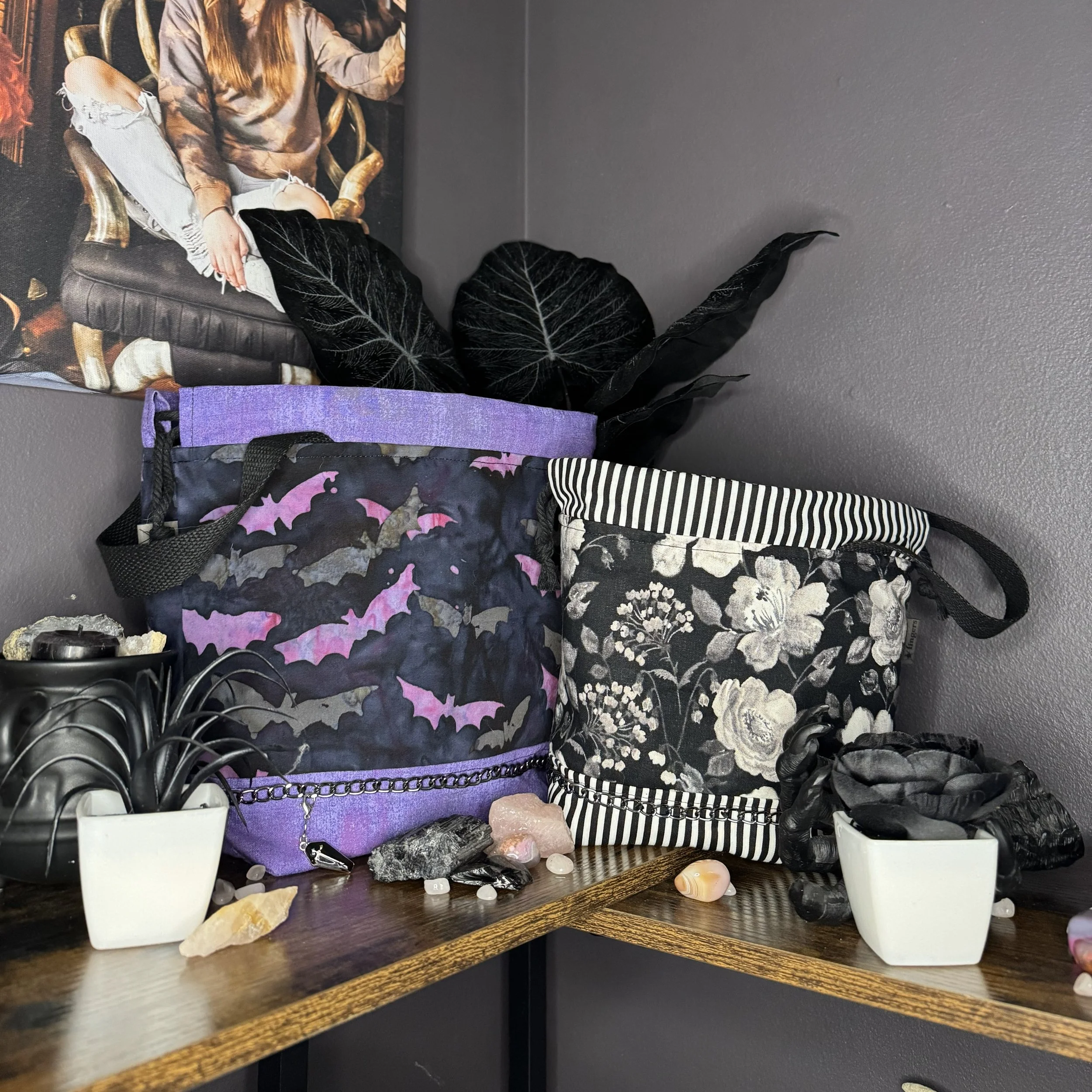 Decorative bags with bat and floral patterns, black gloves, potted plants, and assorted crystals and stones on a wooden shelf against a dark gray wall.