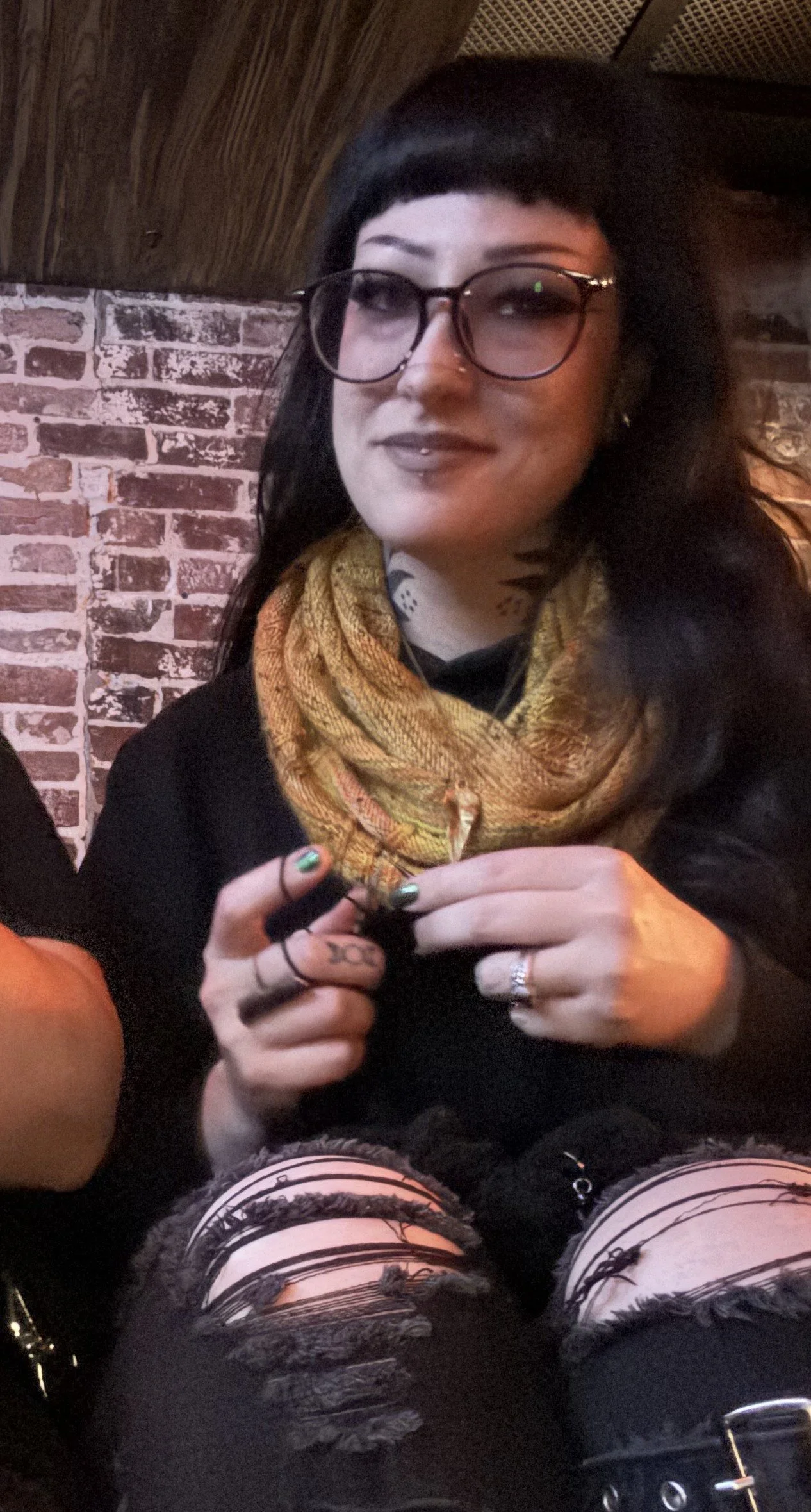A woman with black hair, glasses, and a nose piercing, sitting against a brick wall, wearing a yellow scarf, black top, and ripped black jeans, holding a small object in her hands.