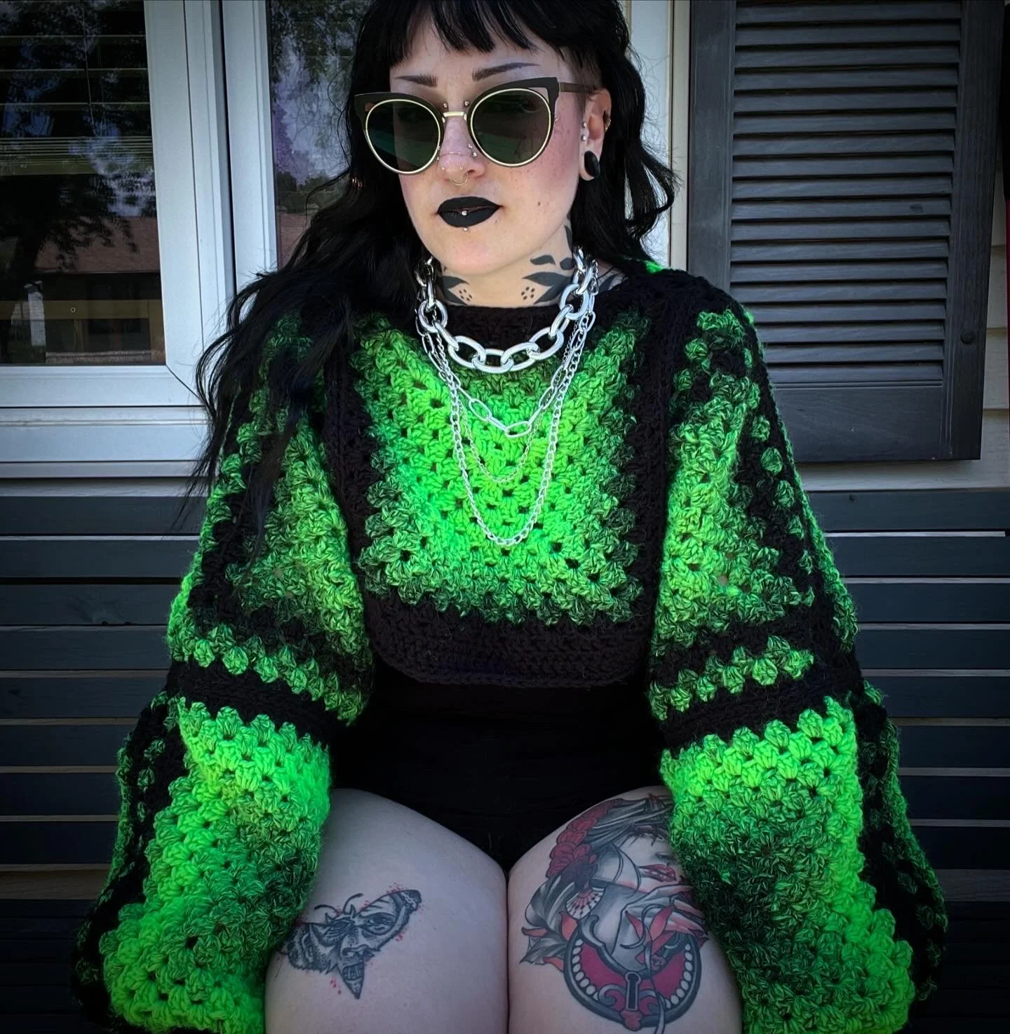 A woman with black hair, sunglasses, and dark makeup sitting outdoors, wearing a bright green and black crocheted sweater, multiple silver necklaces, and tattoos on her thighs.