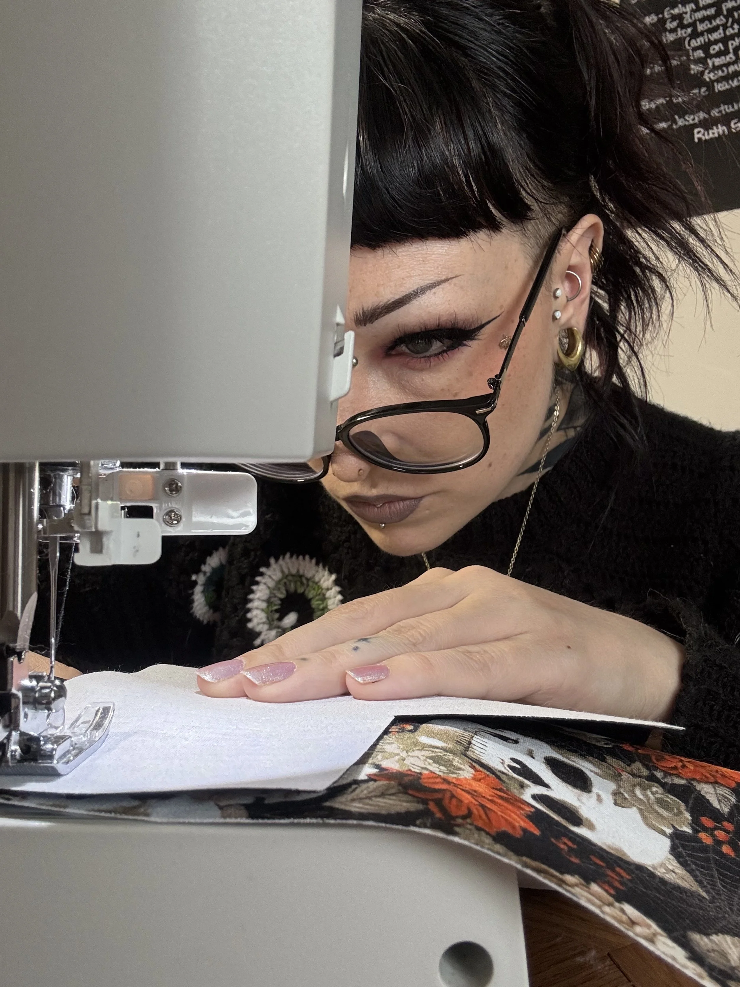 A woman with dark hair and glasses is sewing fabric on a sewing machine, looking closely at her work.