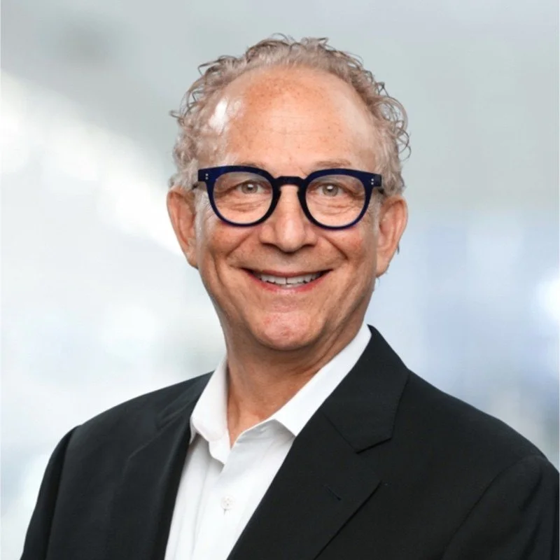 A smiling middle-aged man with curly gray hair, glasses, and a suit against a light, cloudy background.
