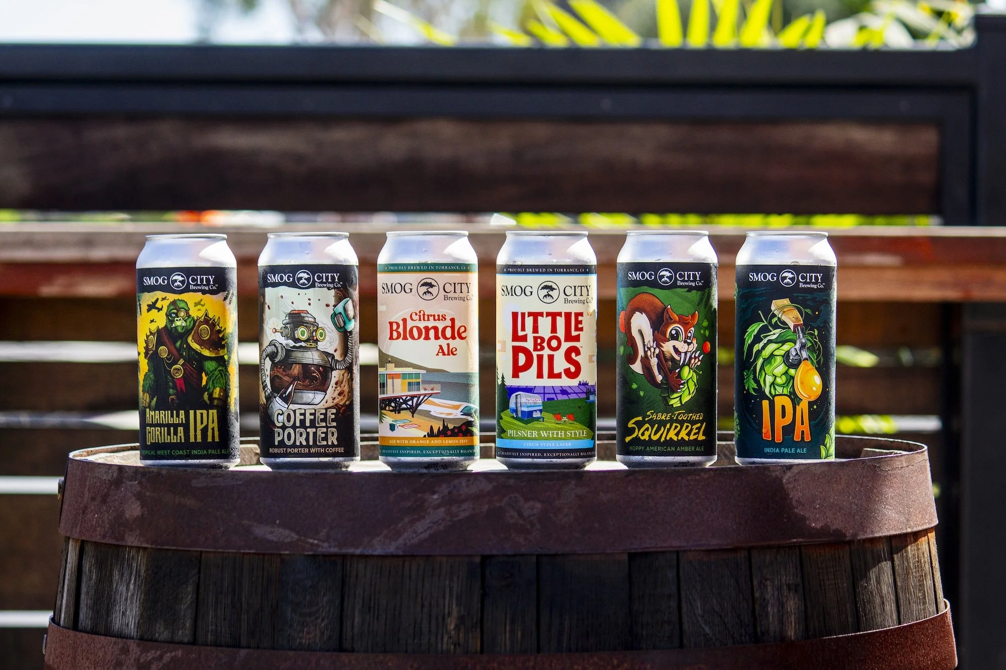 Six colorful cans of craft beers from Smog City Brewing Co., lined up on a wooden barrel outdoors with a wooden fence and green plants in the background.