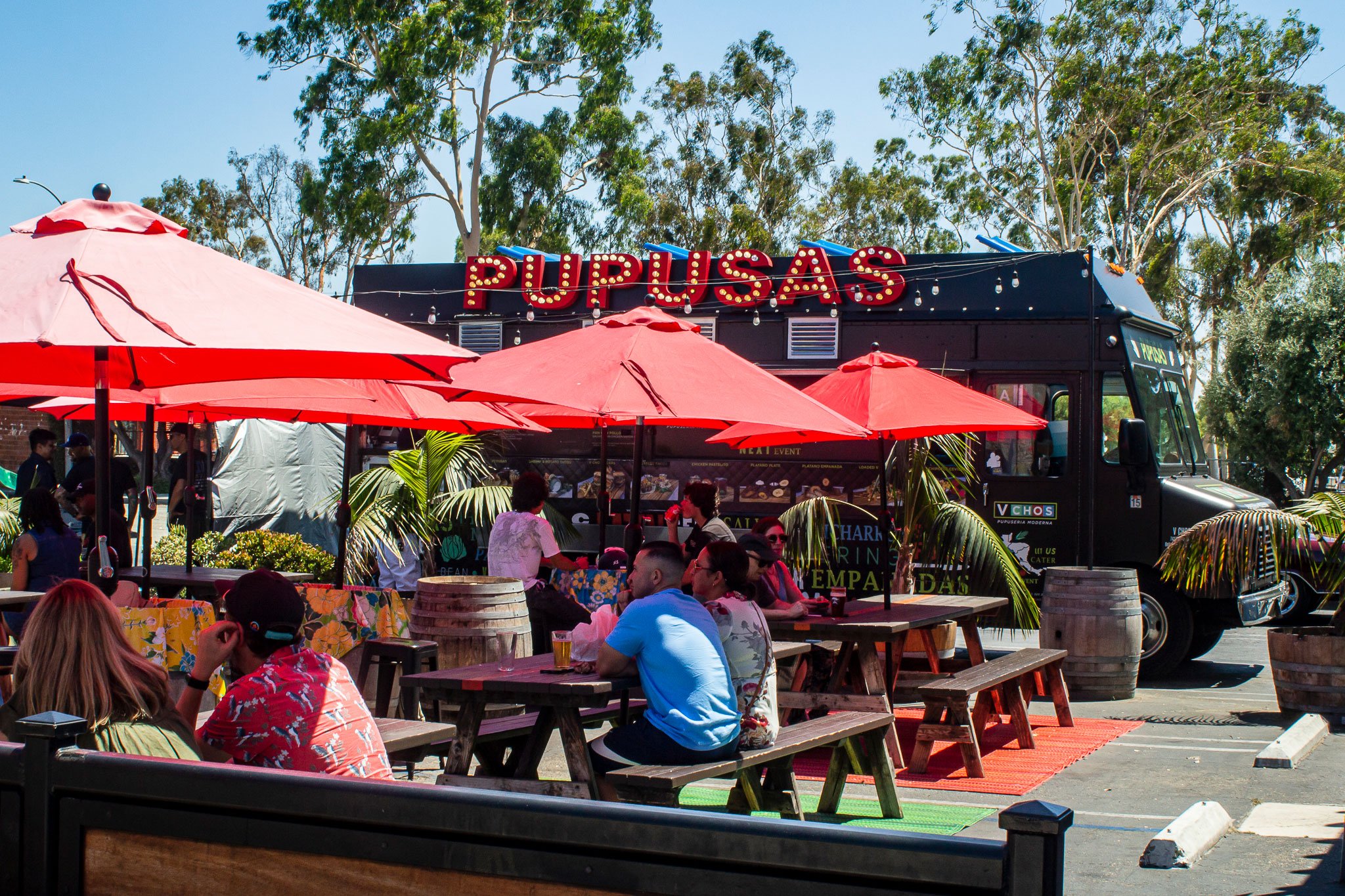 Customers sitting at outside picnic tables under umbrellas with a pupusa food truck in the background.