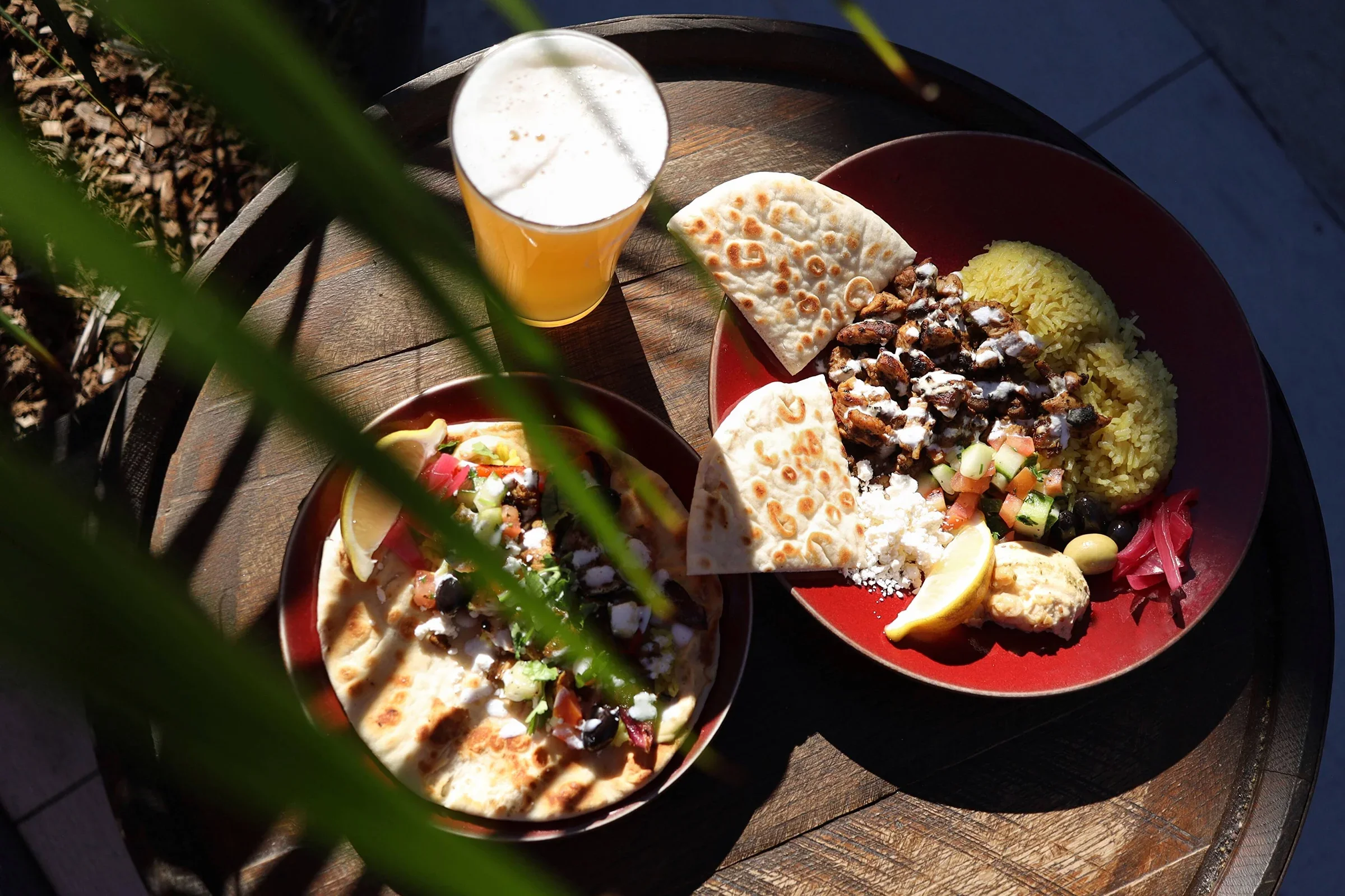 Two plates of Mexican food with rice, beans, meat, tortillas, vegetables, lemon wedges, and a glass of beer on a wooden table.