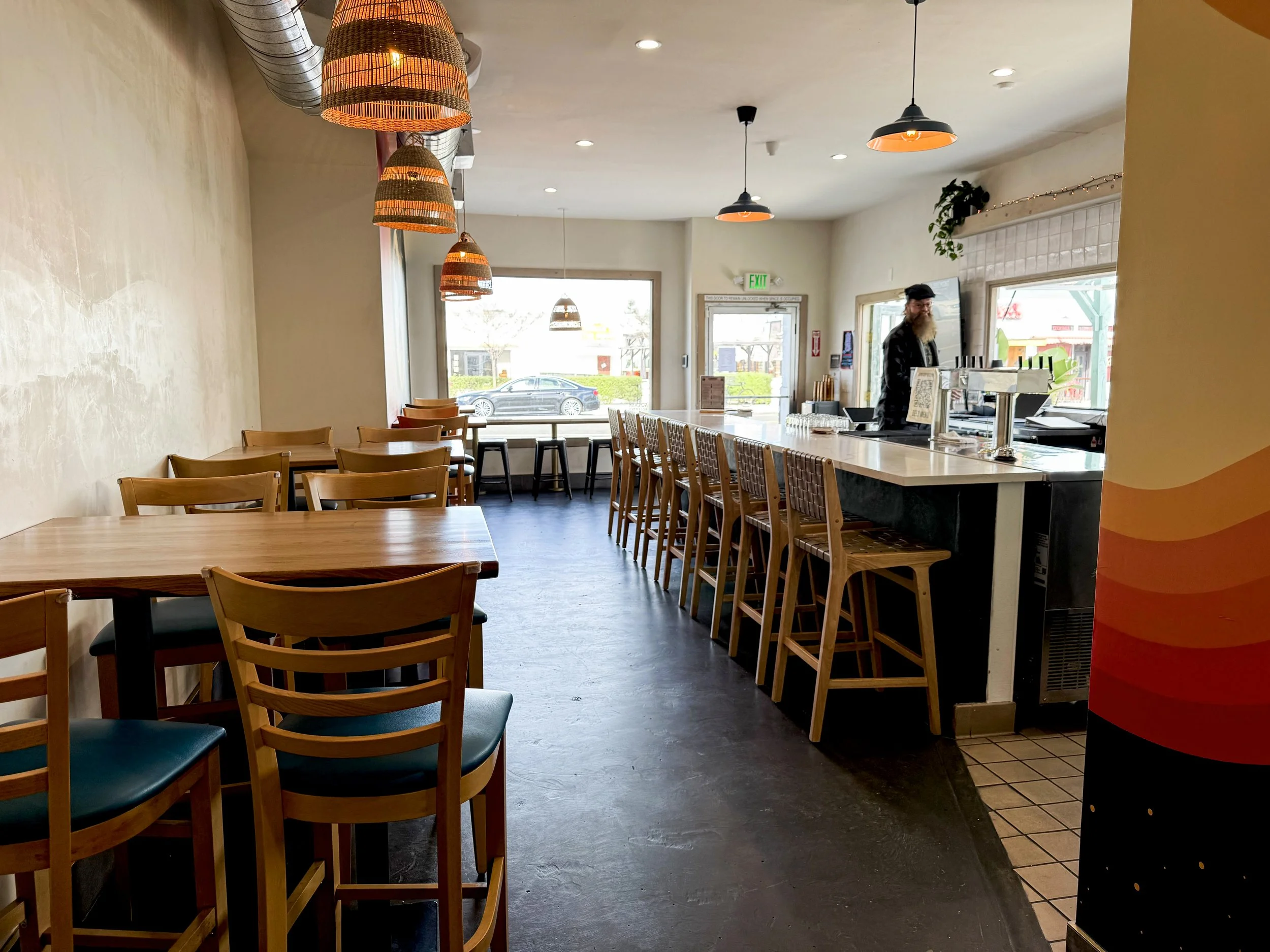 Interior of a coffee shop with a bar counter on the right, barstools, and wooden tables and chairs on the left side. Hanging pendant lights and a large window at the back let in natural light.