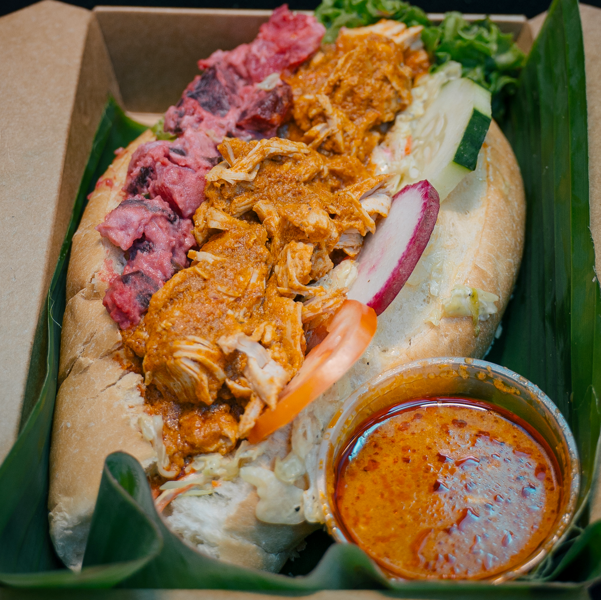 A Vietnamese banh mi sandwich with various meats and vegetables, served with a small cup of spicy sauce.
