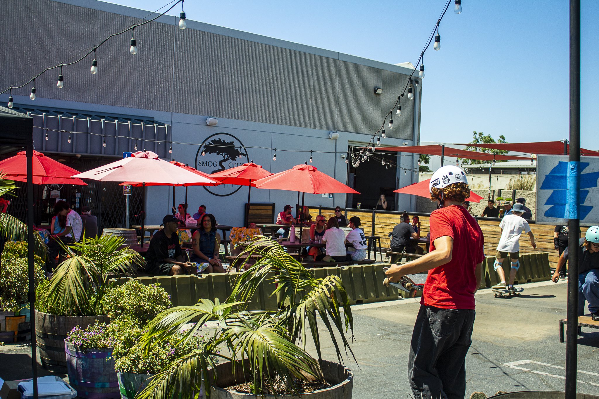 People sitting under red umbrellas and skateboarding in an outdoor area with potted plants and a building with a logo that reads "Smog City".