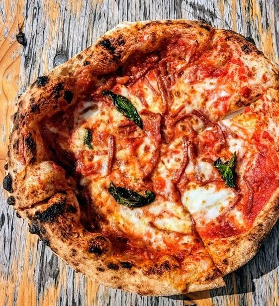 Close-up of a classic Margherita pizza with melted cheese, tomato sauce, basil leaves, and a slightly charred crust on a wooden surface.