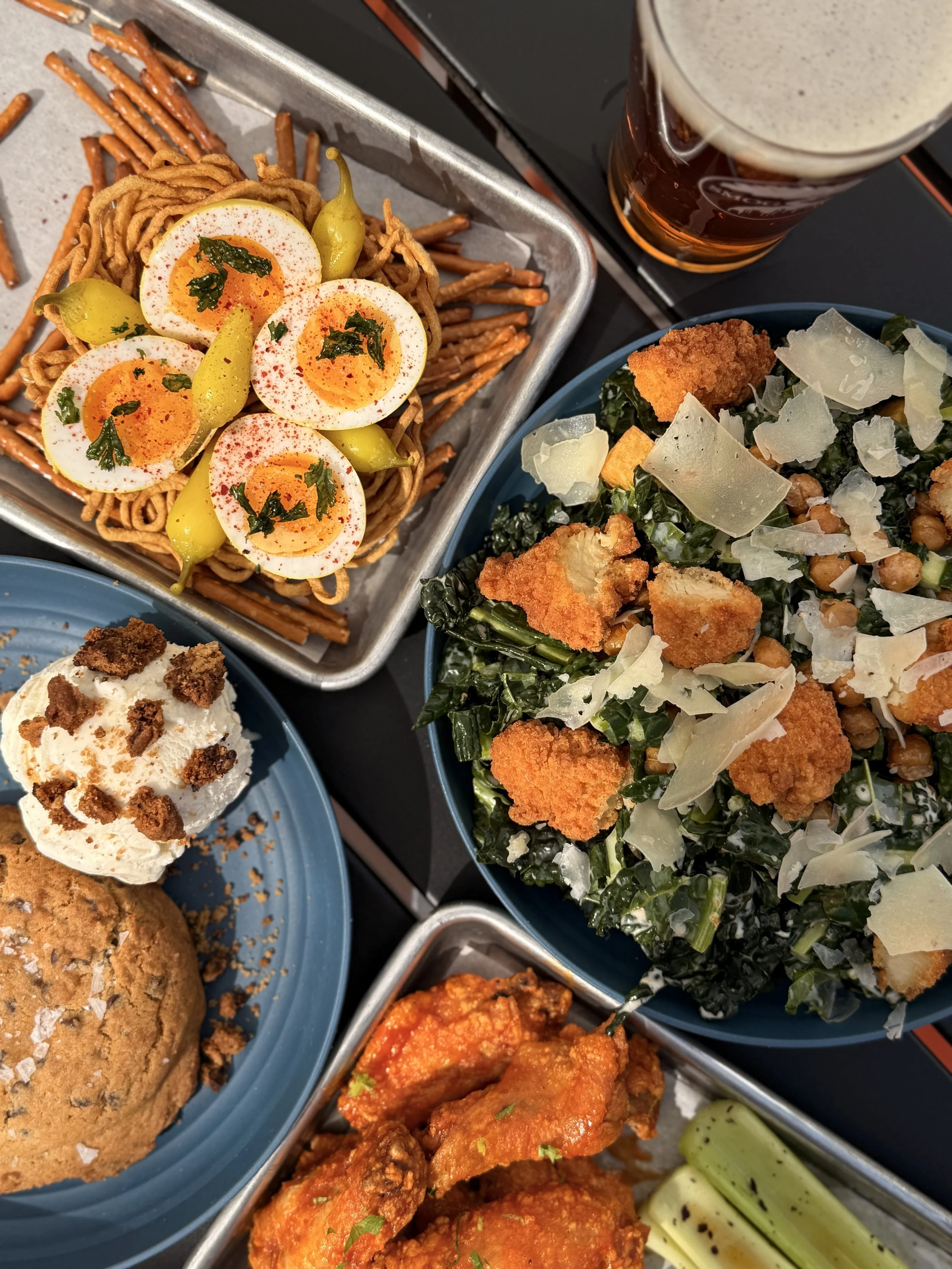 Overhead view of many plates of food, including chicken wings, kale salad, pickled eggs, a cookie, and a pint of beer.