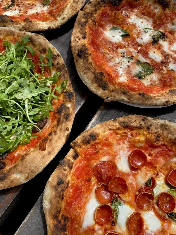 Three personal pizzas with various toppings on a dark wooden surface. One has arugula, another has basil and cheese, and the third has pepperoni slices.