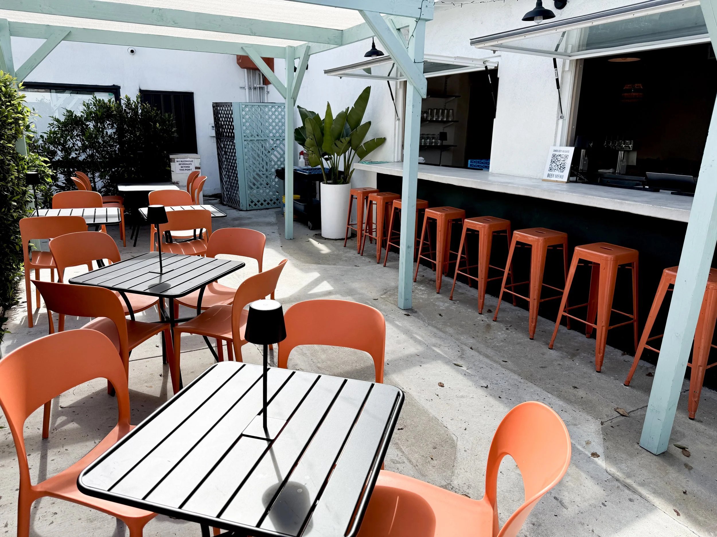 Empty outdoor patio with orange chairs and black and white striped tables, bar stools along a counter, and a white planter with green plants under a light blue pergola.