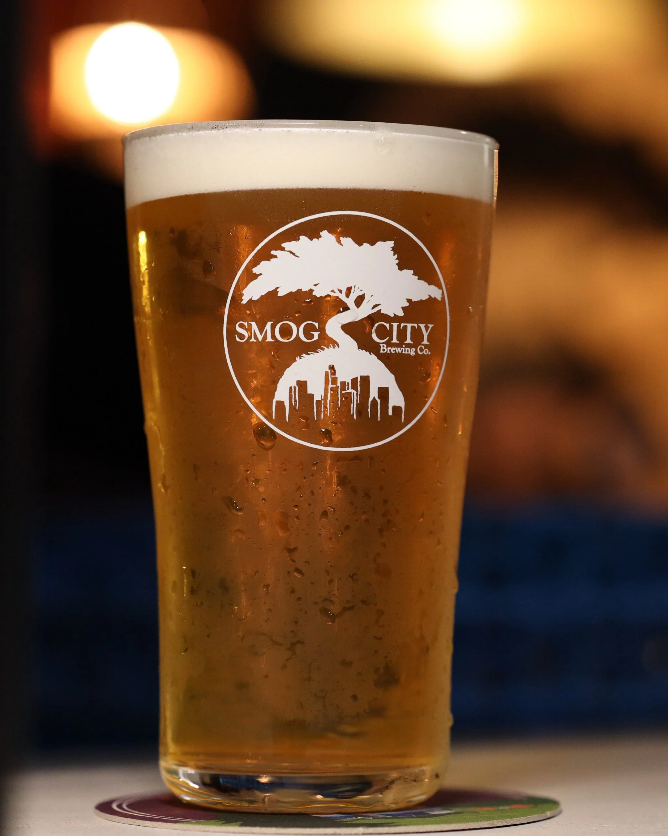 A glass of beer with condensation and foam at the top, with a logo for Smog City Brewing Co. on the glass. The background is blurred with warm lighting.