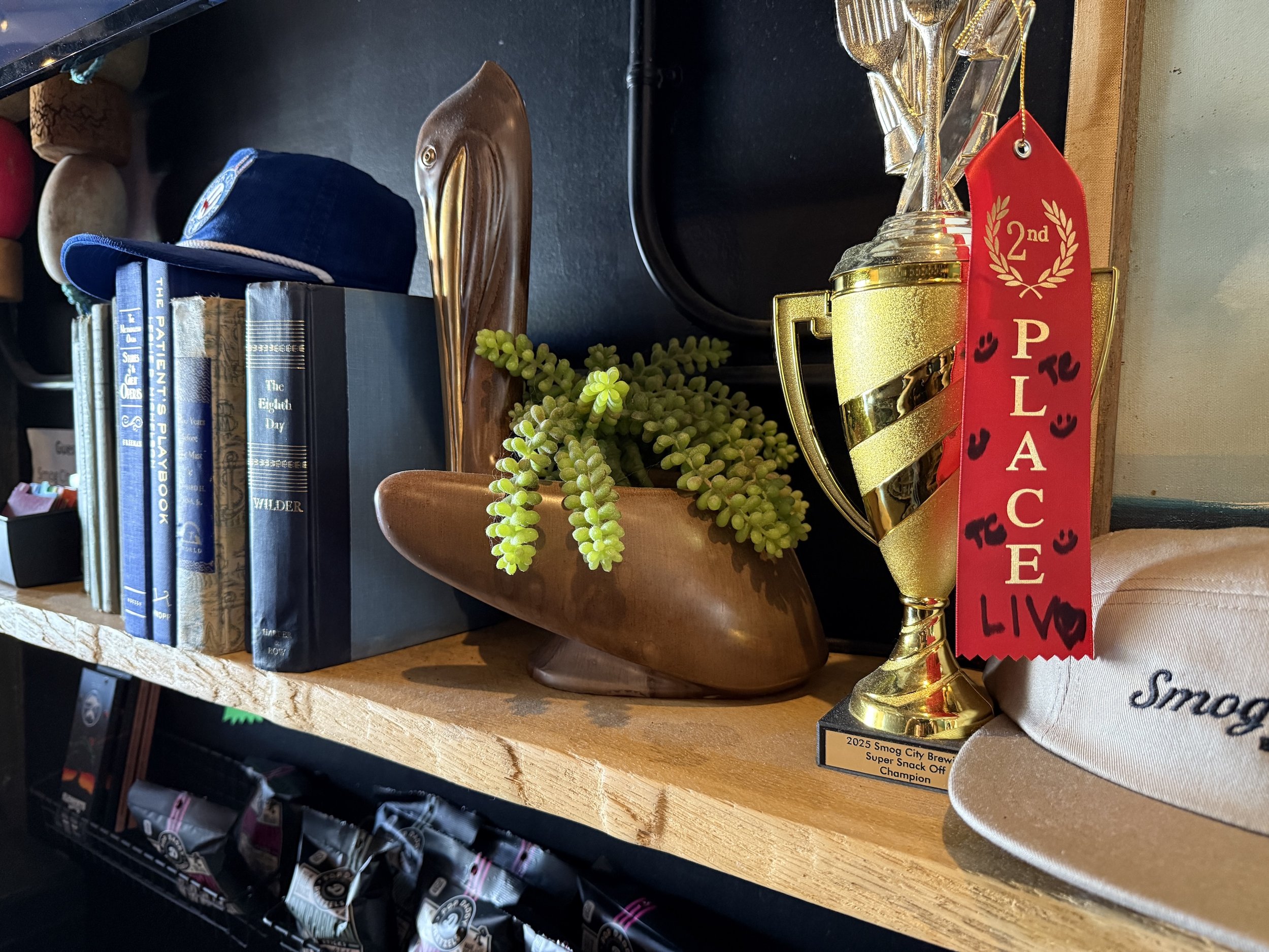 Shelf with books, a blue cap, a wooden decorative object, a plant in a wooden bowl, a trophy with a red ribbon that says '2nd Place', and a beige hat with text.