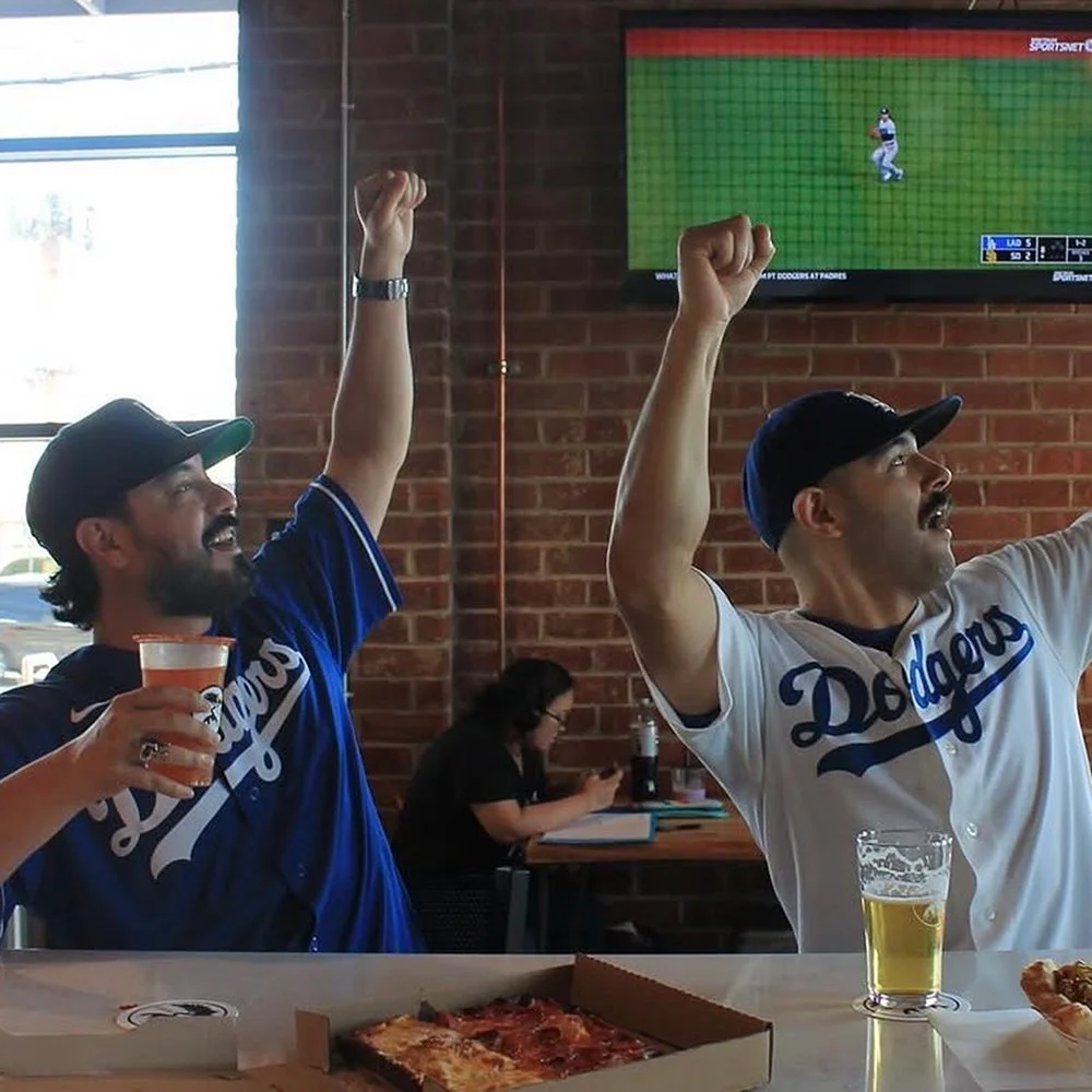 Two customers sitting at a bar with beer and food cheering on a baseball game on television.