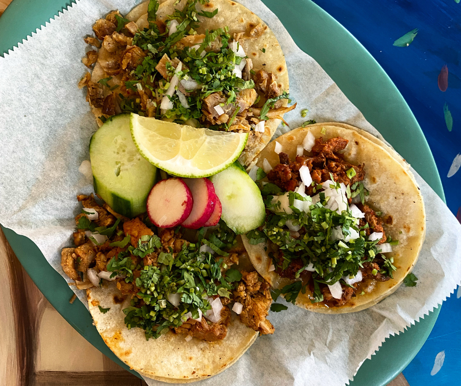 Three tacos on a green plate with lime wedge, cucumber slices, and radish slices, garnished with chopped cilantro and onions.
