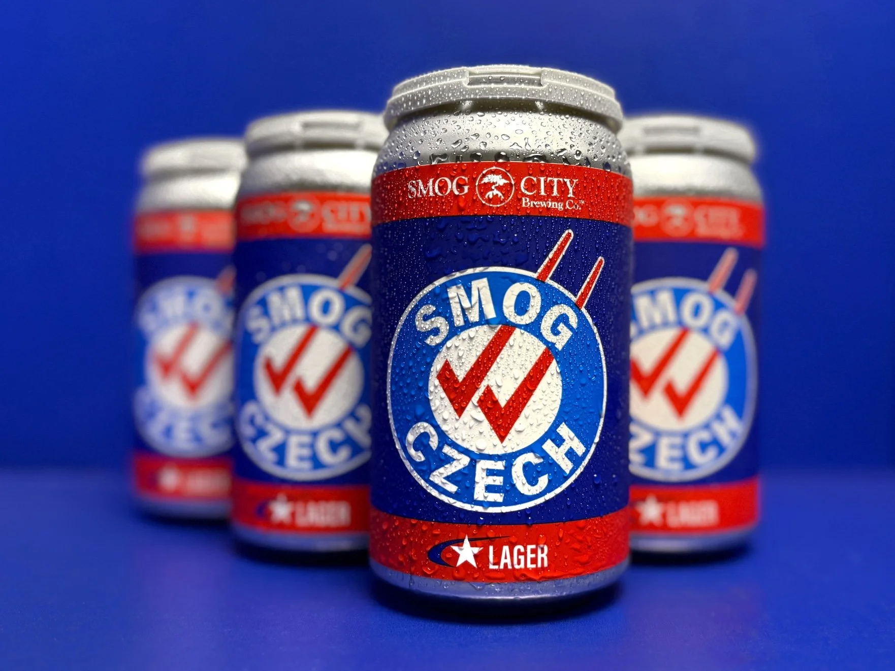 Four cans of Smog Czech beer with condensation droplets, front can prominently displayed, blue background.