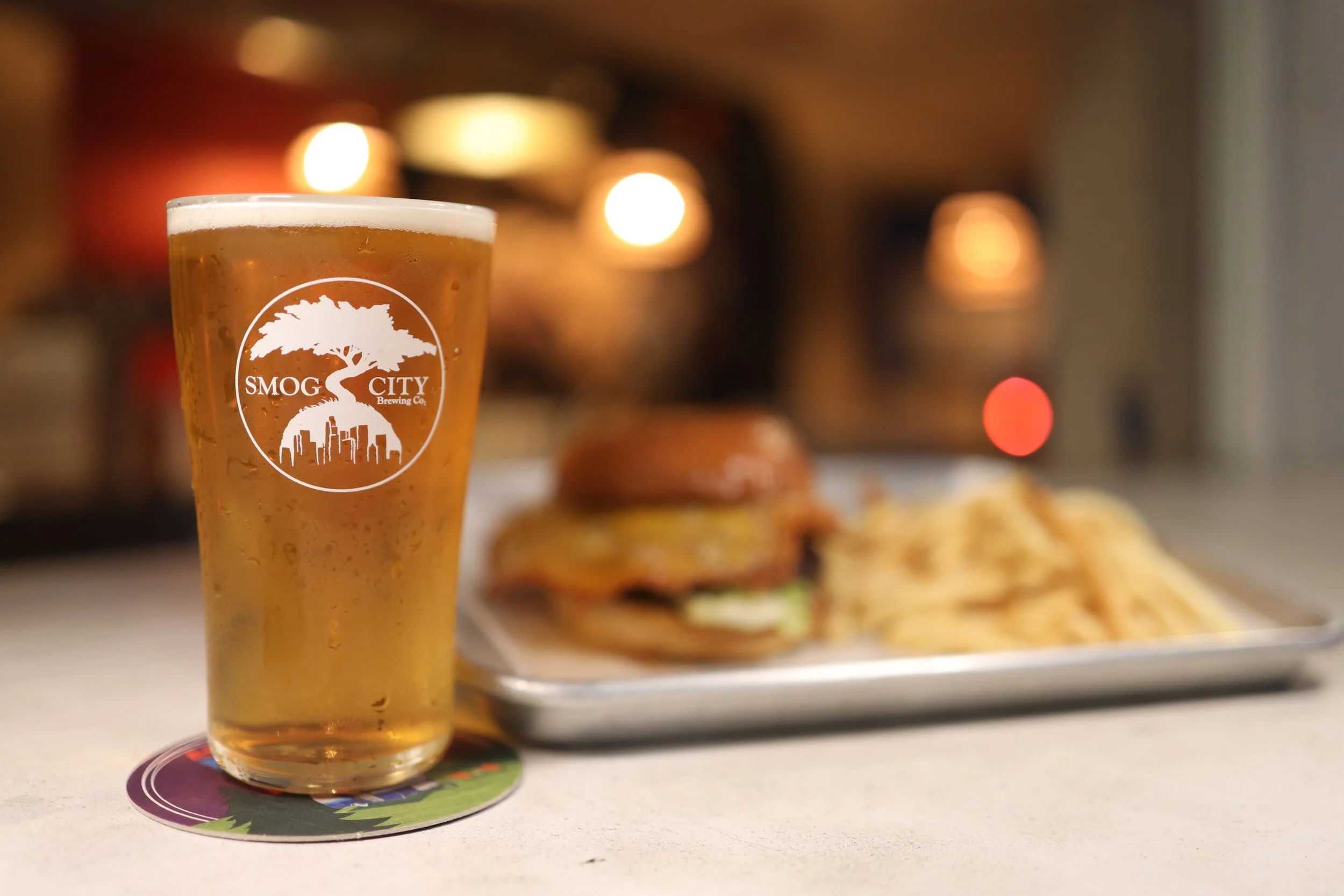 Full pint glass of beer in the foreground with a cheeseburger and fries on the counter in the background.