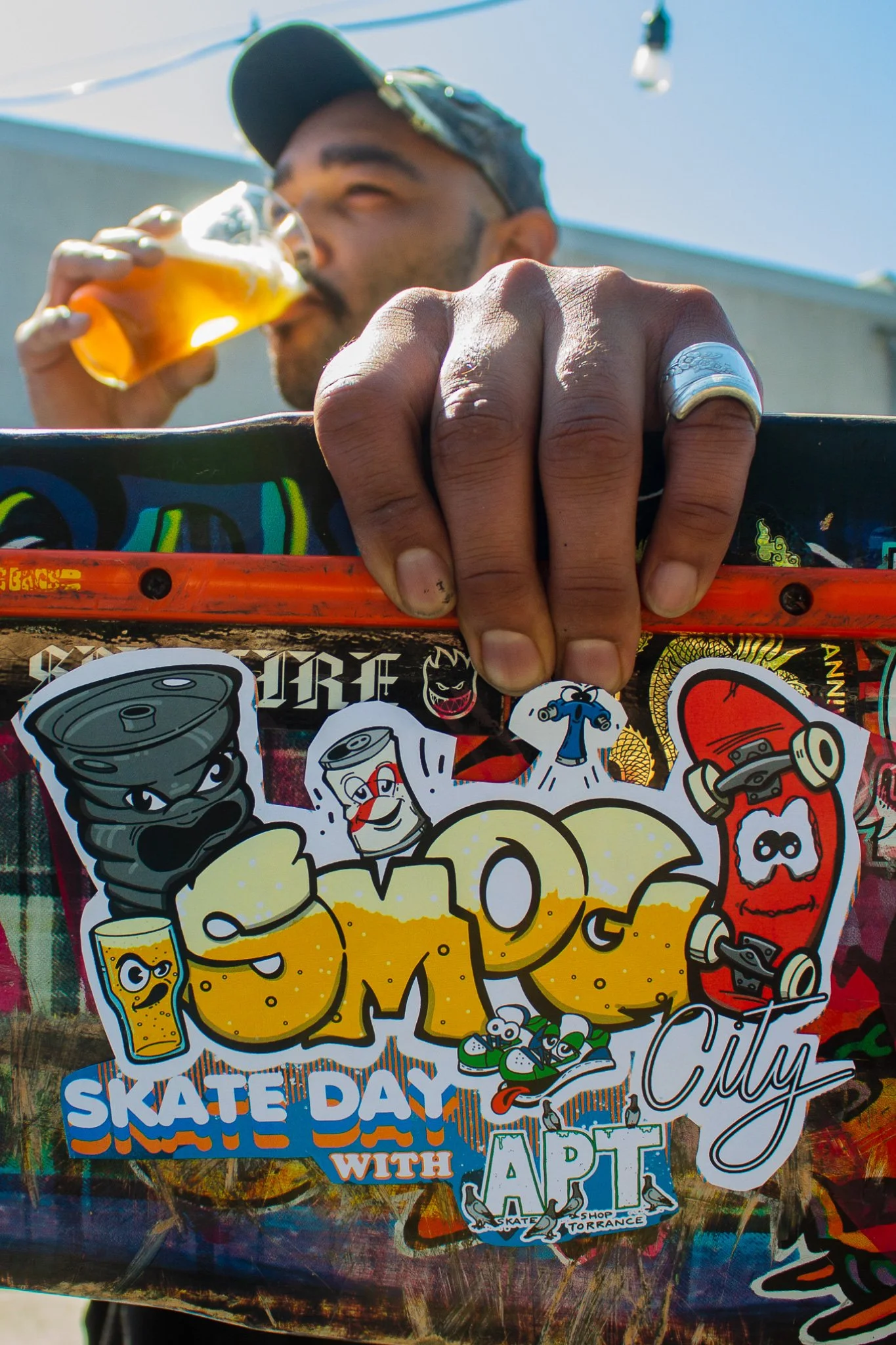 Skateboarder drinking a beer and showing the underside of a skateboard with a Smog City beer sticker.