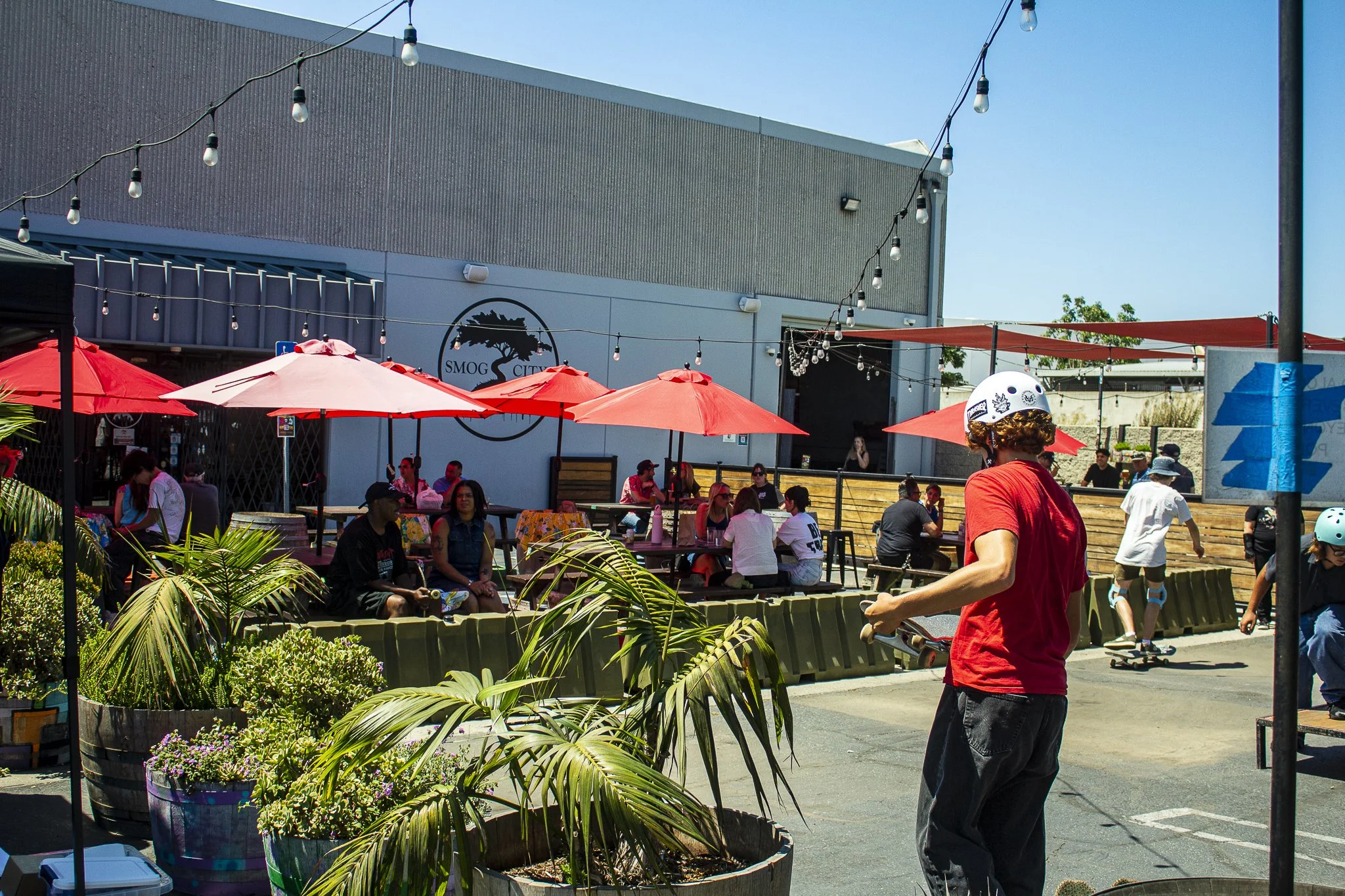 People gathered at an outdoor skate park with a seating area covered by red umbrellas. A person in a red shirt and helmet skates in the foreground, while others skate and sit at tables. There are string lights hanging overhead and a building with a logo that reads 'SMOG CITY' in the background.
