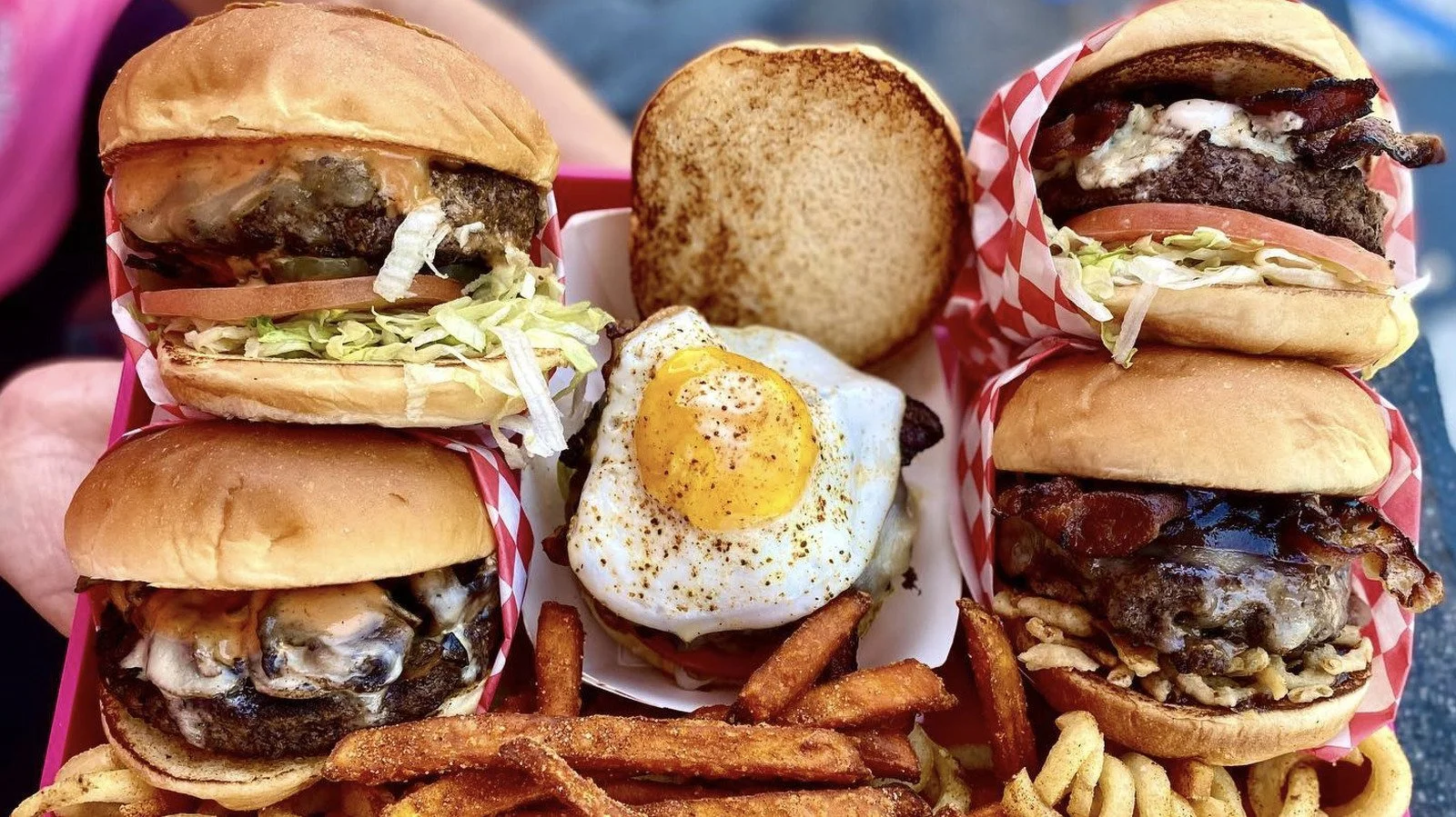 A variety of cheeseburgers with lettuce, tomato, and cheese, accompanied by a fried egg on a bed of fries, and a slice of bread, all in a red and white checkered paper lining.